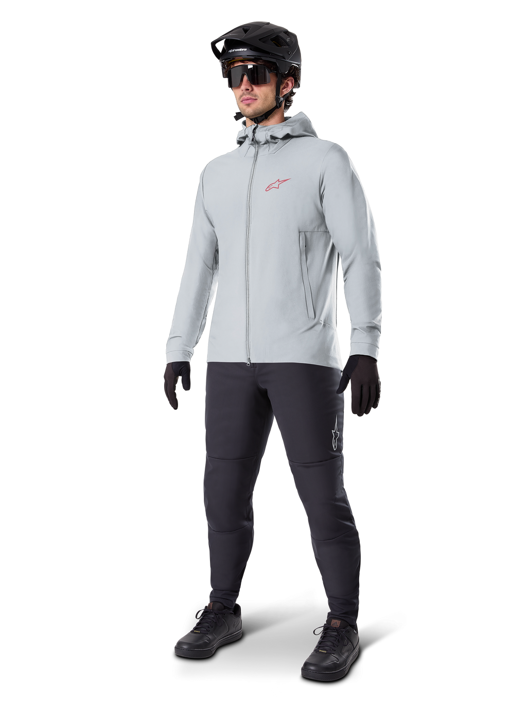 Alpinestars A-Dura Thermal Jacket in Titanium, lichtgrijze fietsjas met capuchon, rood Alpinestars-logo op de borst, gedragen door een man met een zwarte MTB-helm, zonnebril, donkergrijze broek en zwarte handschoenen, professionele mountainbike-uitrusting