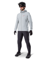 Alpinestars A-Dura Thermal Jacket in Titanium, lichtgrijze fietsjas met capuchon, rood Alpinestars-logo op de borst, gedragen door een man met een zwarte MTB-helm, zonnebril, donkergrijze broek en zwarte handschoenen, professionele mountainbike-uitrusting
