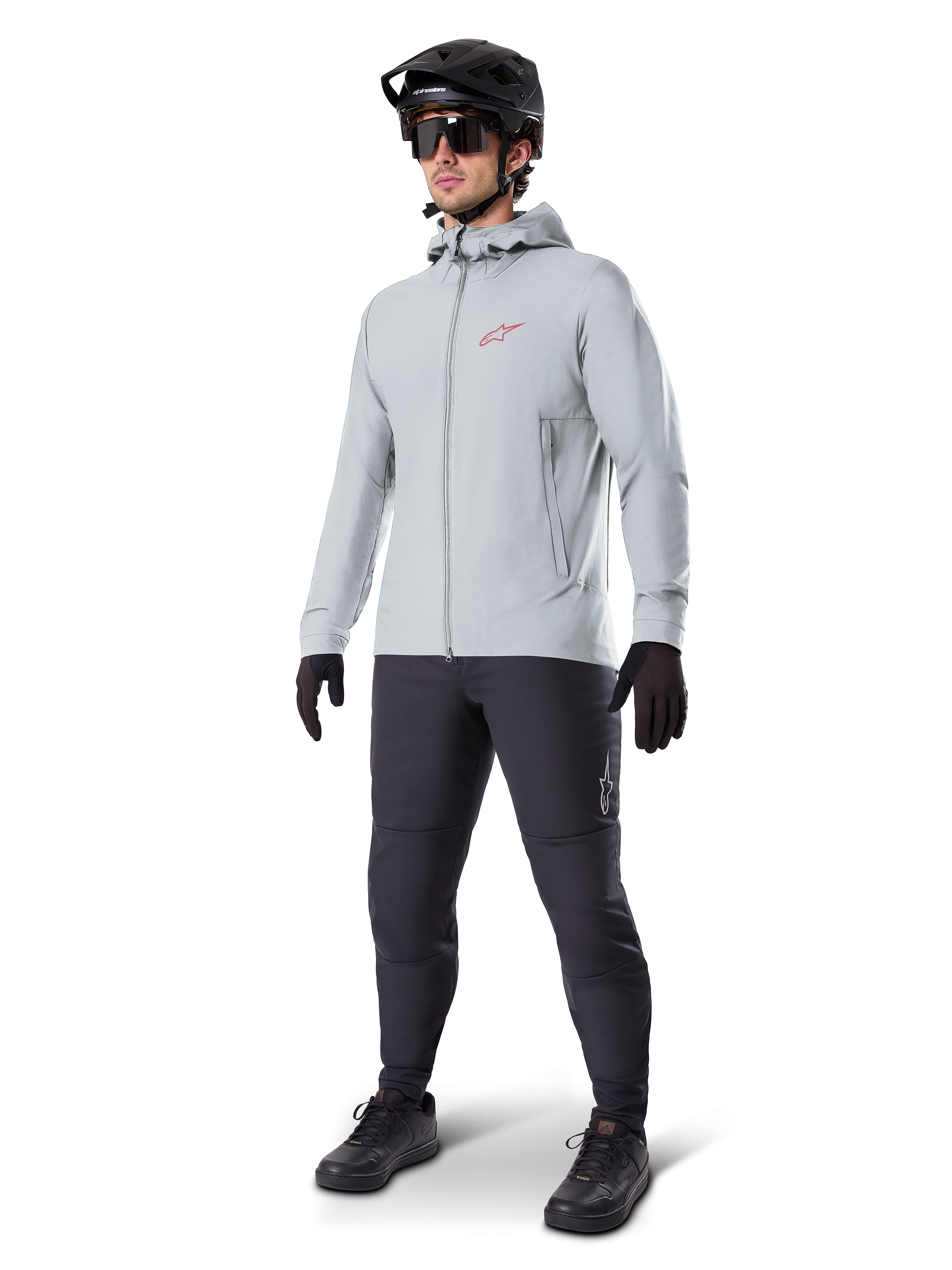 Alpinestars A-Dura Thermal Jacket in Titanium, lichtgrijze fietsjas met capuchon, rood Alpinestars-logo op de borst, gedragen door een man met een zwarte MTB-helm, zonnebril, donkergrijze broek en zwarte handschoenen, professionele mountainbike-uitrusting