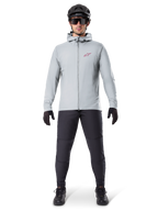 Alpinestars A-Dura Thermal Jacket, titanium grijs, fietsjas gedragen met zwarte mountainbikebroek, handschoenen en helm, voorzien van een klein rood sterlogo op de borst en een volledige ritssluiting aan de voorzijde, lichtgewicht geïsoleerde kleding voor off-road ritten
