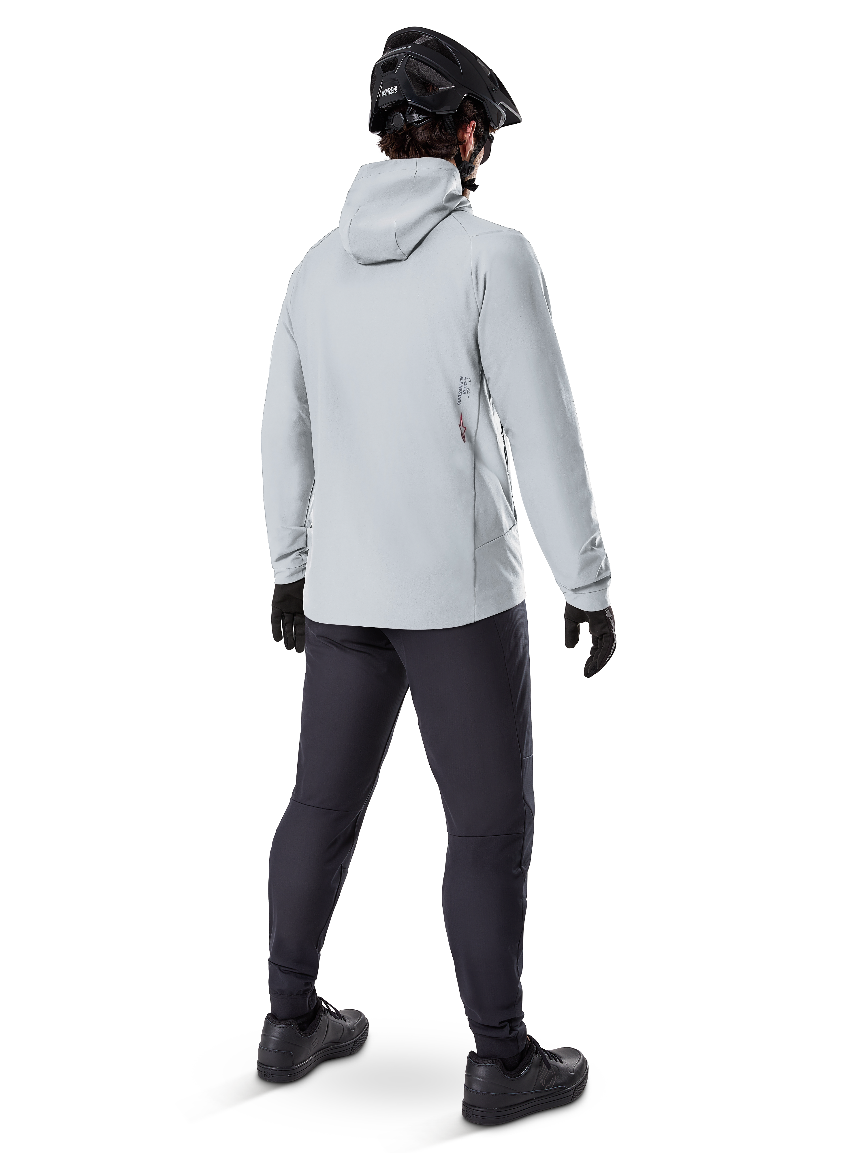 Alpinestars A-Dura Thermal Jacket in Titanium, achteraanzicht van mannelijke fietser die een lichtgrijze thermische jas met capuchon draagt, gecombineerd met zwarte MTB-helm, zwarte handschoenen en donkergrijze broek, functionele mountainbike-uitrusting