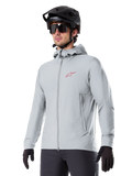 Alpinestars A-Dura Thermal Jacket, lichtgrijze geïsoleerde fietsjas, model met capuchon en een klein rood logo, gedragen met een zwarte helm en handschoenen, technische mountainbikekleding voor ritten in koud weer