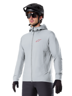 Alpinestars A-Dura Thermal Jacket, lichtgrijze geïsoleerde fietsjas, model met capuchon en een klein rood logo, gedragen met een zwarte helm en handschoenen, technische mountainbikekleding voor ritten in koud weer