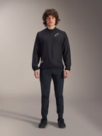 Modèle portant la veste coupe-vent Alpinestars A-Aria en noir, dotée d'une fermeture éclair intégrale, d'un col montant et d'un logo Astars blanc sur la poitrine, associée à un pantalon technique noir et des chaussures noires, vêtement de cyclisme performant pour le VTT et l'extérieur