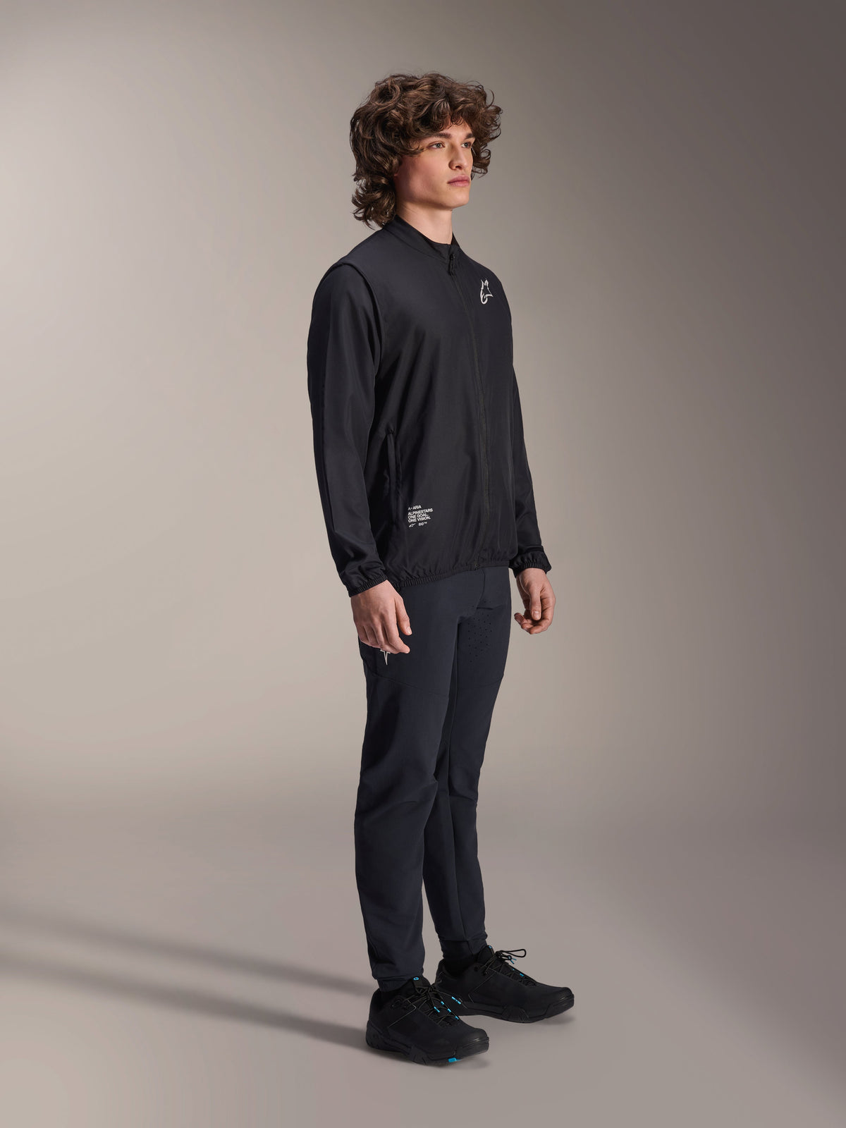 Alpinestars A-Aria Windbreaker Jack, zwart, lichtgewicht windbestendig fietsjack gedragen door een model met krullend haar, met een wit Astars-logo op de borst en branding rechtsonder, gecombineerd met een zwarte broek en schoenen, prestatiegerichte pasvorm voor mountainbiken en gravelrijden