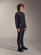 Veste coupe-vent Alpinestars A-Aria, noire, veste de cyclisme légère et résistante au vent portée par un mannequin aux cheveux bouclés, avec un logo Astars blanc sur la poitrine et un marquage en bas à droite, associée à un pantalon et des chaussures noirs, coupe performance pour le VTT et le gravel