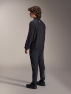 Veste coupe-vent Alpinestars A-Aria, noire, veste de cyclisme légère et résistante au vent, vue de trois-quarts arrière sur modèle, dotée d'un petit logo blanc au niveau de la nuque et d'un ourlet élastiqué, associée à un pantalon et des chaussures noirs assortis pour un look axé sur la performance.