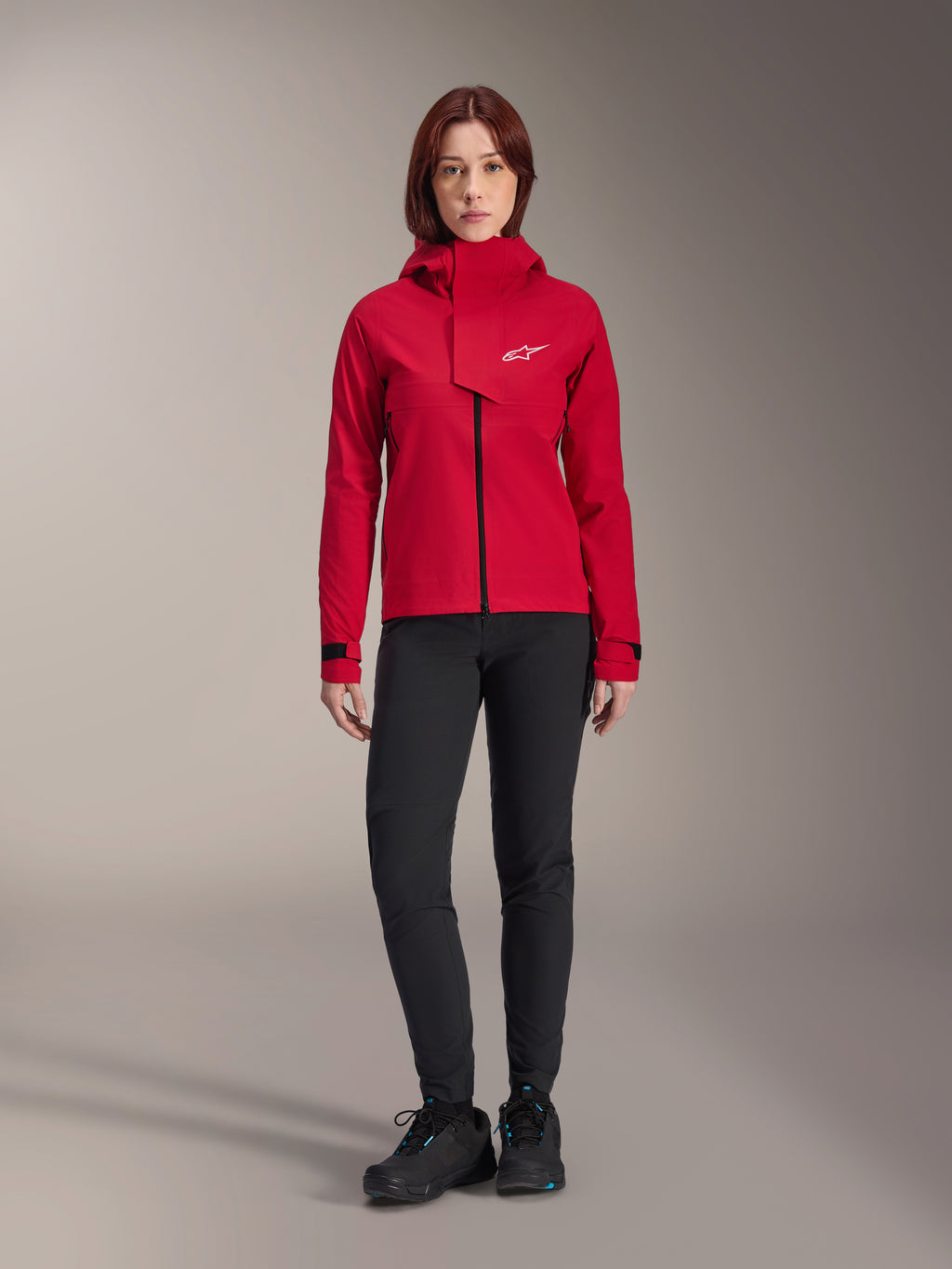 Alpinestars Stella A-Dura Elite Waterdichte Jas, MTB-jas voor dames, Rood Fluo, gedragen met een zwarte broek en schoenen, voorzien van een capuchon met hoge hals en wit Alpinestars-logo, technische mountainbike-kleding