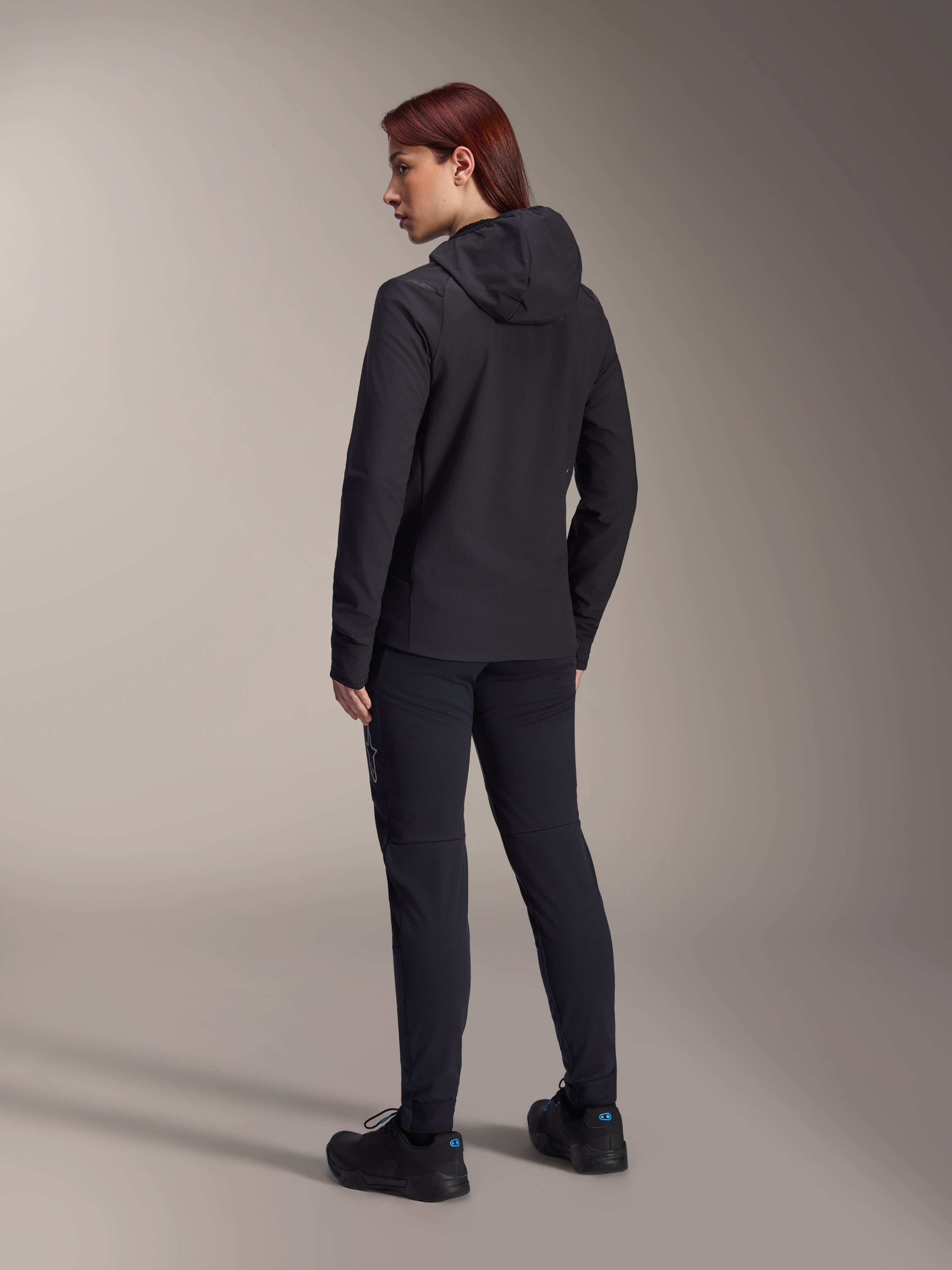 Alpinestars Stella A-Dura Thermal Jacket, MTB-fietsjack voor dames, zwart, achteraanzicht met focus op de geïsoleerde capuchon en normale pasvorm, gedragen met bijpassende zwarte broek en schoenen, lichtgewicht thermische isolatie voor ritten in koud weer