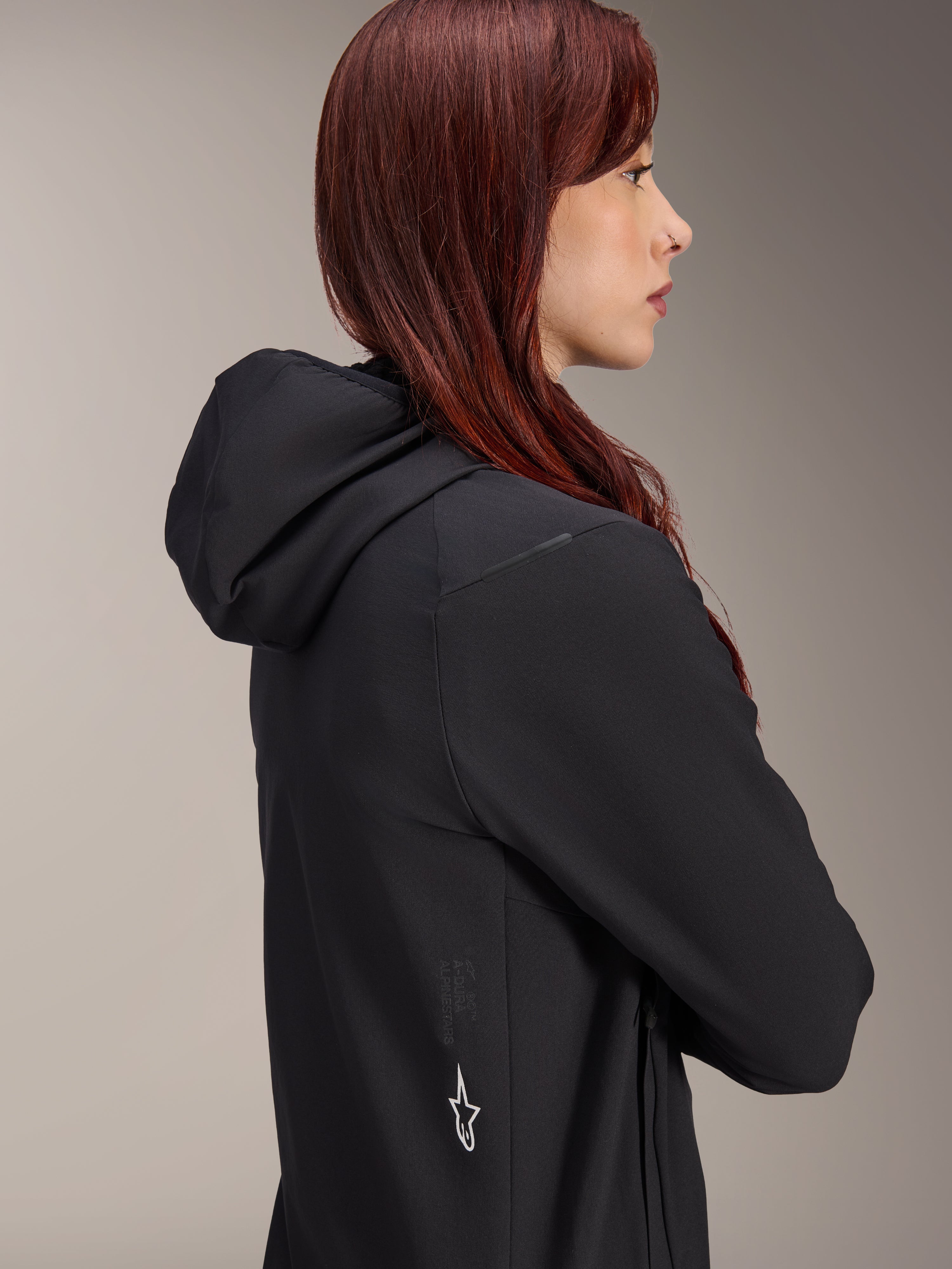 Alpinestars Stella A-Dura Thermal Jacket, dames fietsjack, zwart, zijprofiel van een vrouw met rood haar die de jas draagt, voorzien van een geïntegreerde capuchon en wit Alpinestars sterlogo aan de zijkant, lichtgewicht geïsoleerde thermische kleding voor MTB