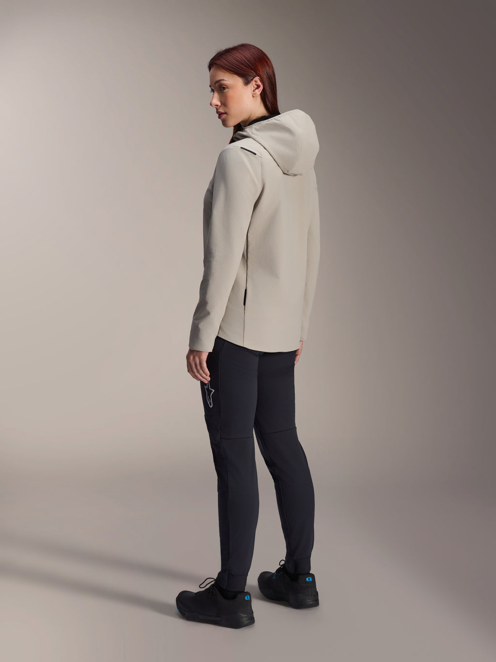 Alpinestars Stella A-Dura Thermal Jacket en gris Titanium, vue de trois-quarts arrière d'une femme portant la veste de cyclisme isolée à capuche et un pantalon de VTT noir, photo en studio montrant la coupe régulière et les détails des poches.