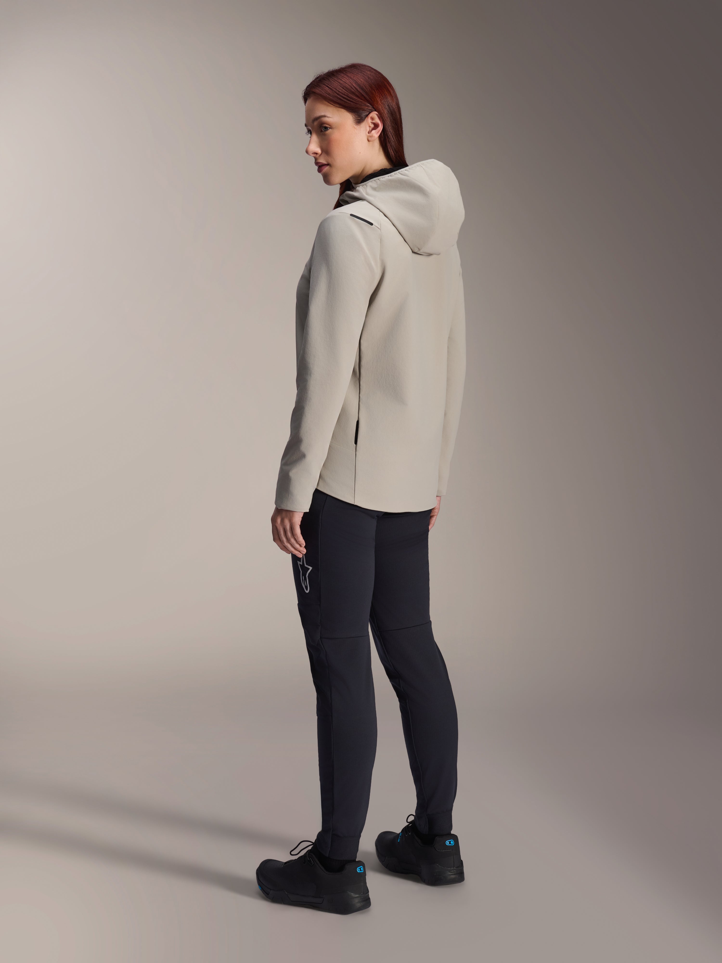 Alpinestars Stella A-Dura Thermal Jacket en gris Titanium, vue de trois-quarts arrière d'une femme portant la veste de cyclisme isolée à capuche et un pantalon de VTT noir, photo en studio montrant la coupe régulière et les détails des poches.