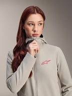 Veste thermique Stella A-Dura Alpinestars pour femme, gris titane avec un logo étoile Alpinestars rose, mannequin aux longs cheveux roux fermant la fermeture éclair de la veste à capuche, vêtement de cyclisme léger et isolant pour temps froid