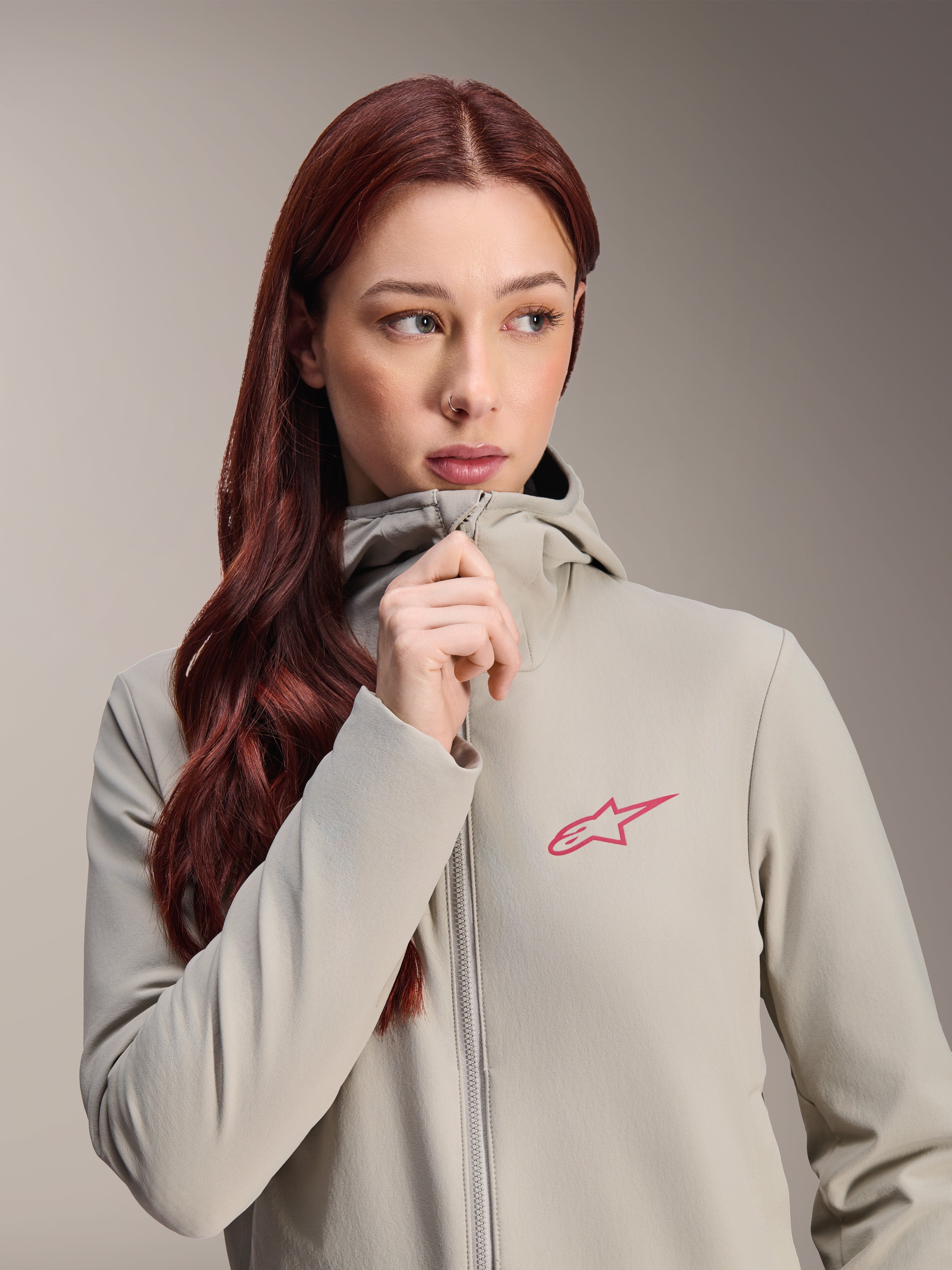 Veste thermique Stella A-Dura Alpinestars pour femme, gris titane avec un logo étoile Alpinestars rose, mannequin aux longs cheveux roux fermant la fermeture éclair de la veste à capuche, vêtement de cyclisme léger et isolant pour temps froid