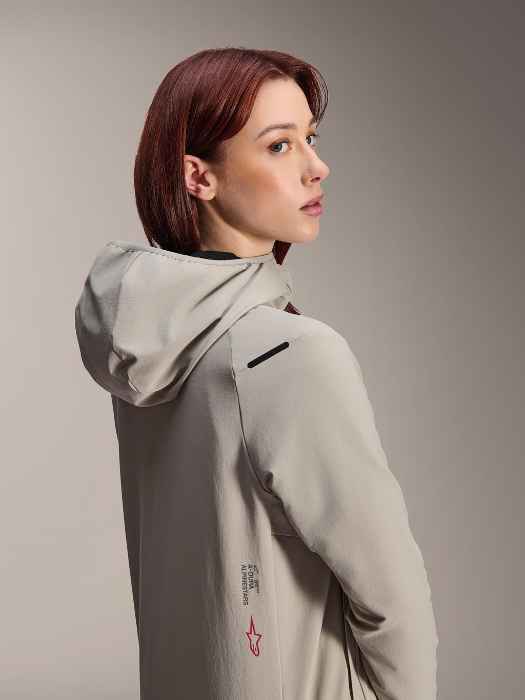 Veste thermique Stella A-Dura Alpinestars, veste de VTT pour femme, gris titane, femme regardant par-dessus son épaule portant une veste technique à capuche, avec inscription Alpinestars et logo étoile rouge sur le côté, conçue pour le cyclisme par temps froid