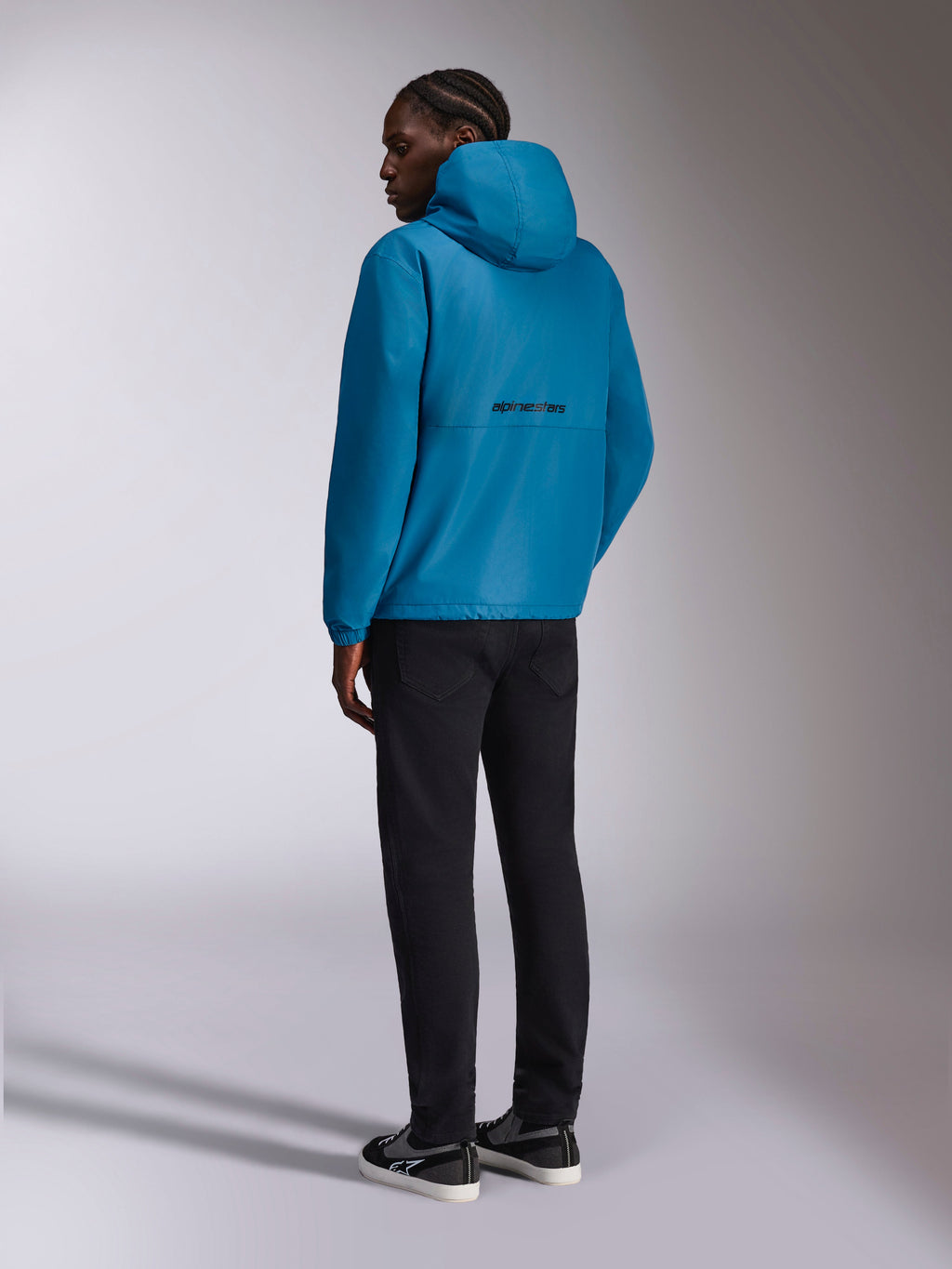 Alpinestars Speeder Chromium Windbreaker Jas, windbreaker met capuchon, blauw, achteraanzicht met zwart Alpinestars-woordlogo op de rug, gedragen met een zwarte broek en zwart-witte sneakers, casual motorsport-geïnspireerde streetwear