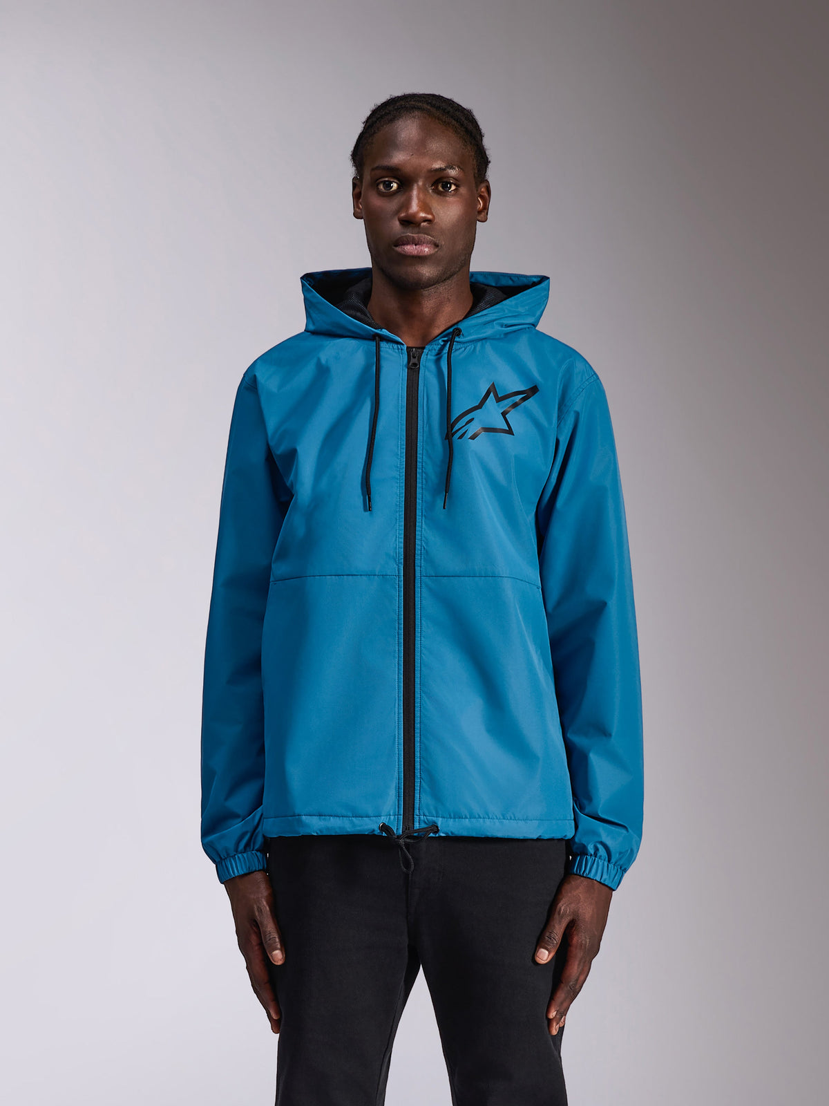 Alpinestars Speeder Chromium Windbreak Jacket, blaue Kapuzen-Windjacke mit schwarzem Astars-Logo auf der Brust, durchgehender Frontreißverschluss, elastische Bündchen und Kapuze mit Kordelzug, von einem Model als lässige Sportbekleidung getragen.