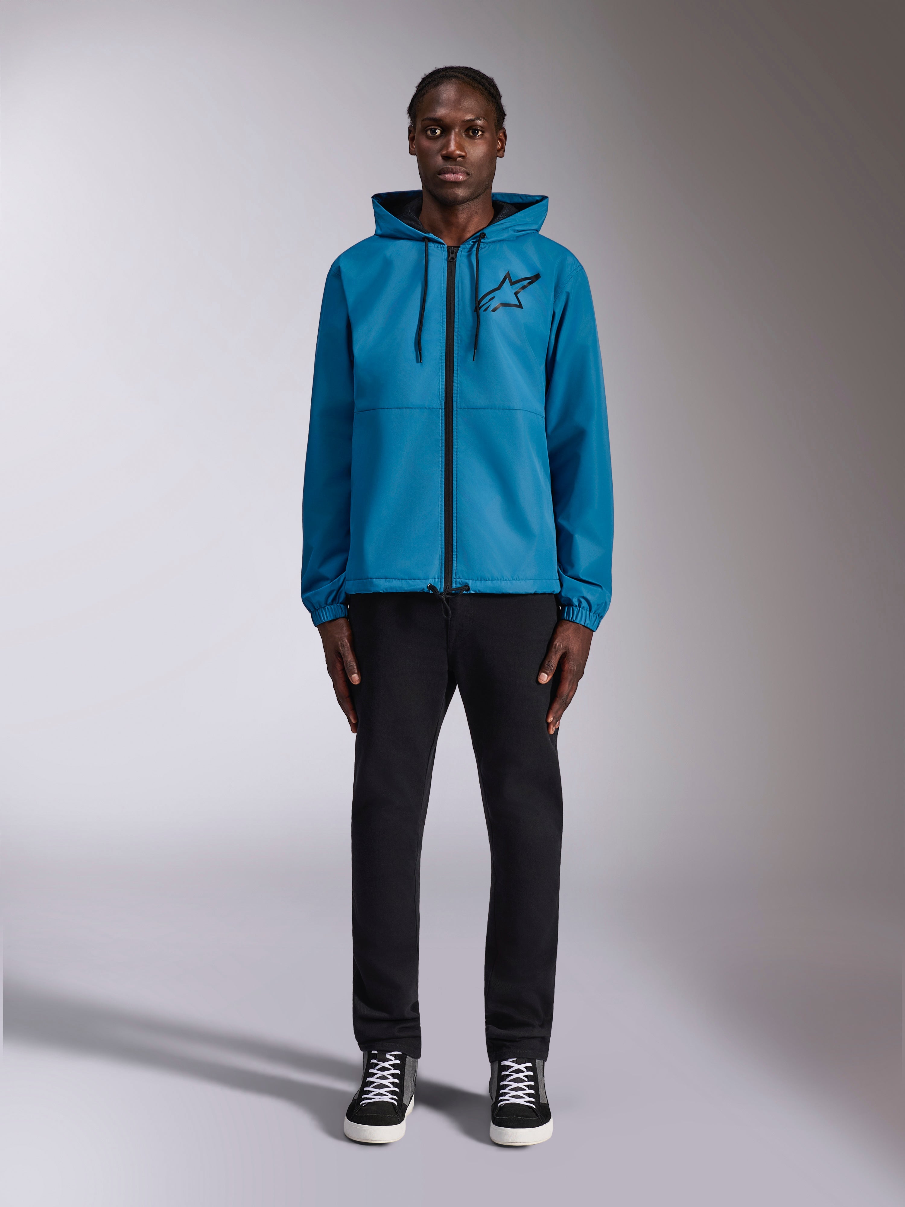 Model draagt de Alpinestars Speeder Chromium Windbreak Jacket in blauw, voorzien van een ontwerp met capuchon, zwarte trekkoorden, een zwarte rits aan de voorkant en een zwart Astars-logo op de borst, gecombineerd met een zwarte broek en sneakers, casual motorsport streetwear