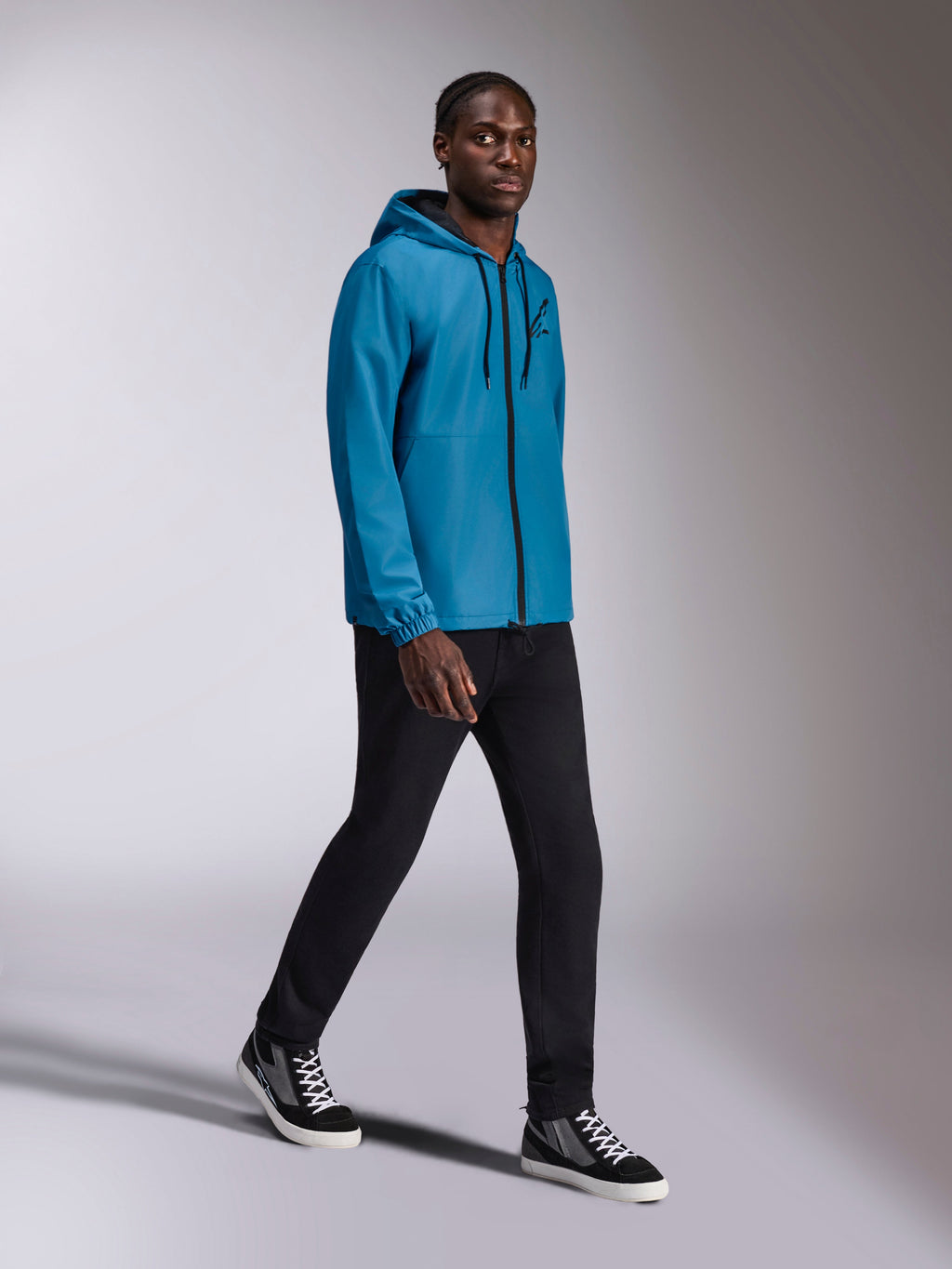 Alpinestars Speeder Chromium Windbreaker Jacket, blauwe windbreaker met capuchon gedragen door een mannelijk model, zwarte rits aan de voorzijde en logo op de borst, gecombineerd met zwarte broek en hoge sneakers, casual op motorsport geïnspireerde sportkleding
