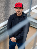 Homme portant la veste Alpinestars Overhaul Racer noire, une veste de pluie à capuche résistante à l'eau avec un petit logo sur la poitrine, accompagnée d'une casquette Alpinestars rouge, vêtement lifestyle motorsport décontracté
