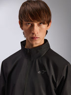 Veste Alpinestars Overhaul Racer, veste de pluie noire, portrait rapproché d'un mannequin portant la veste, avec un col montant et un petit logo Astars argenté sur la poitrine, vêtement d'extérieur décontracté d'inspiration sport mécanique