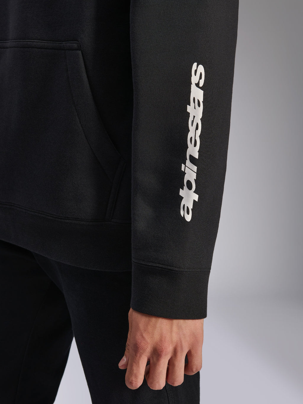 Alpinestars Harken Hoodie, zwart, close-up van de mouw met verticaal wit woordmerklogo, ribboord en kangoeroezak-detail, casual sportkleding fleece