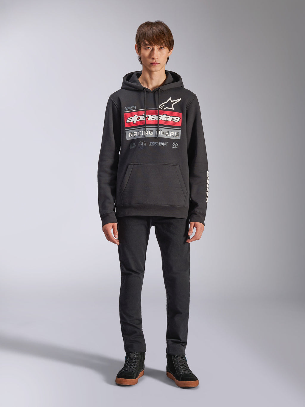 Alpinestars Harken hoodie, zwarte pullover hoodie, opvallende grafische print op de borst met rode strepen en de tekst 'Racing Ahead', wit Astars-logo, kangoeroezak, capuchon met trekkoord, casual motorsport-geïnspireerde streetwear, persoon draagt de hoodie met zwarte broek