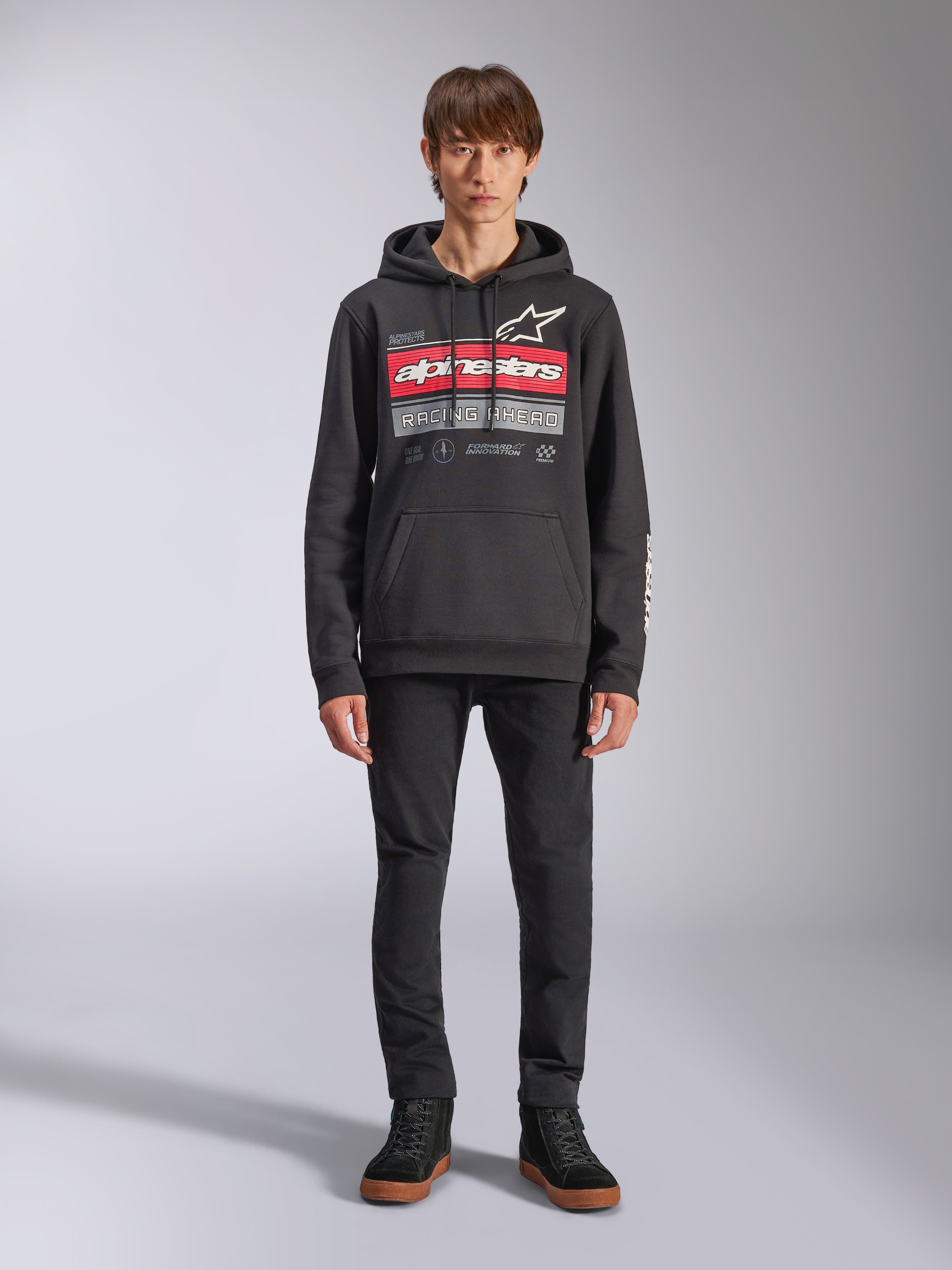 Alpinestars Harken hoodie, zwarte pullover hoodie, opvallende grafische print op de borst met rode strepen en de tekst 'Racing Ahead', wit Astars-logo, kangoeroezak, capuchon met trekkoord, casual motorsport-geïnspireerde streetwear, persoon draagt de hoodie met zwarte broek