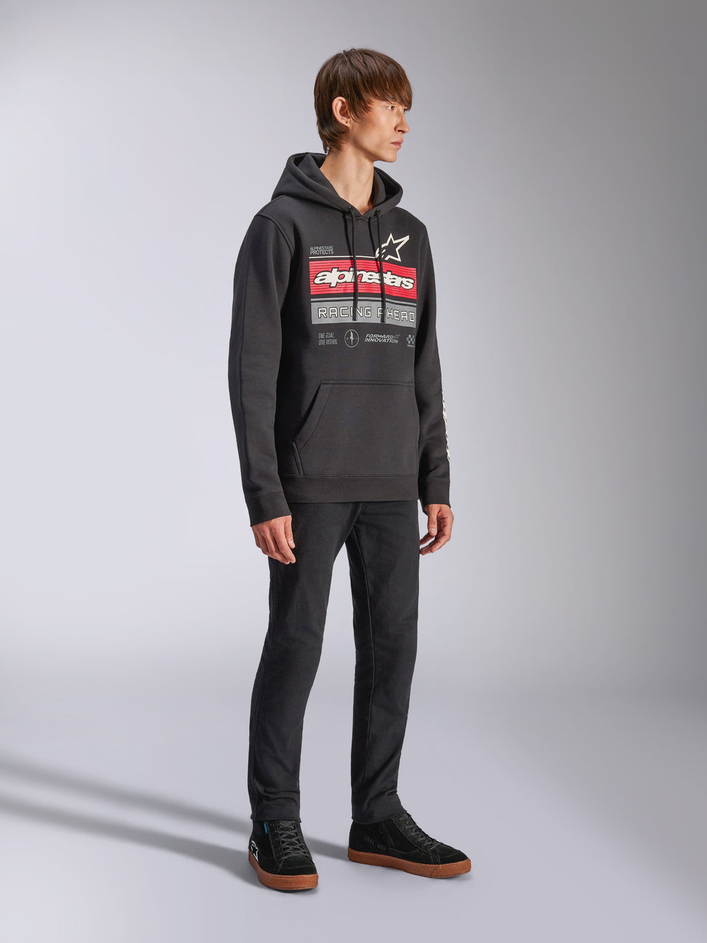 Alpinestars Harken Hoodie, zwarte pullover fleece hoodie met een grote grafische print op de borst, rood Alpinestars woordmerk en sterlogo, kangoeroezak, gedragen door een model met zwarte broek en sneakers, casual motorsport streetwear