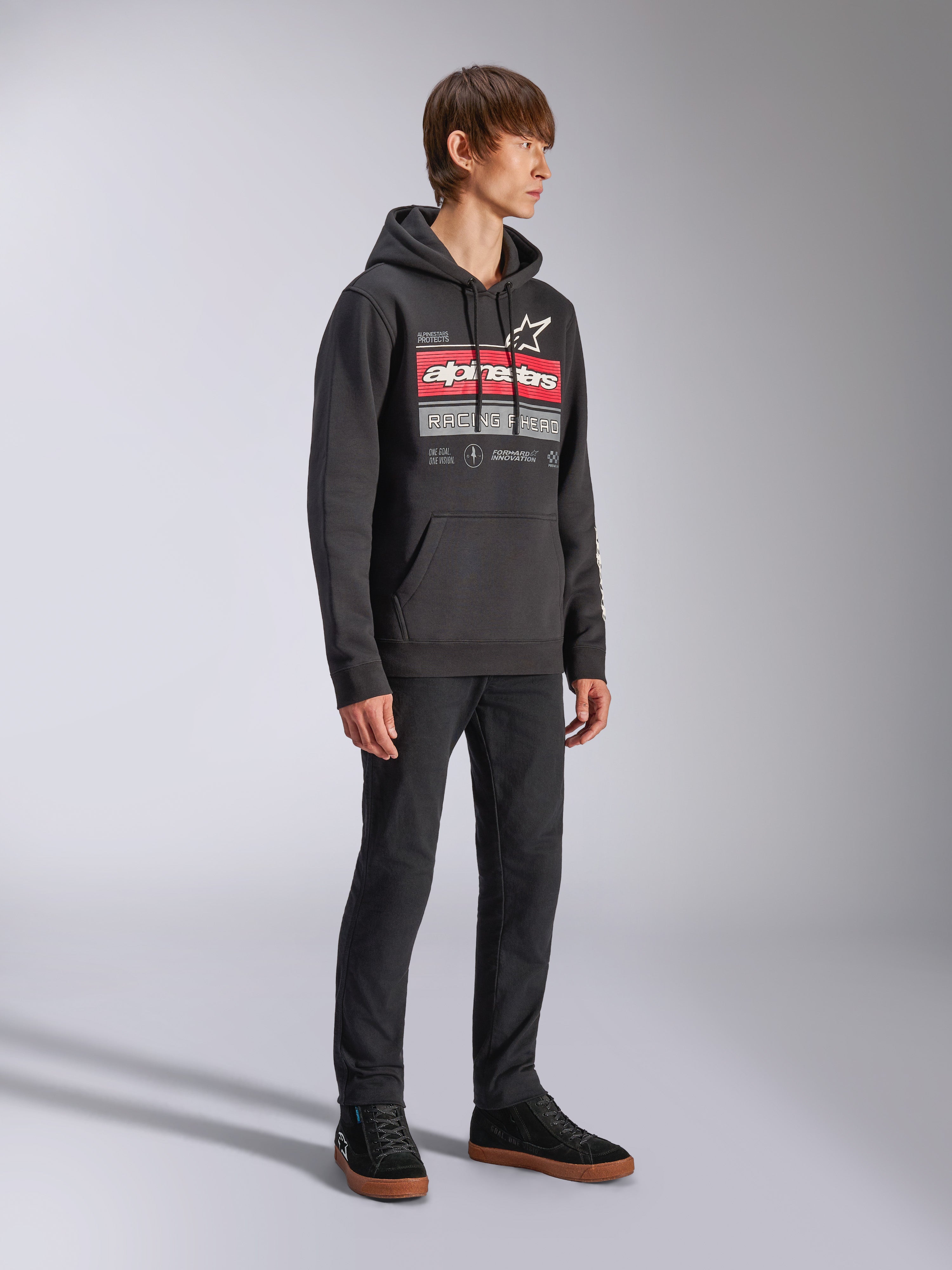 Alpinestars Harken Hoodie, zwarte pullover fleece hoodie met een grote grafische print op de borst, rood Alpinestars woordmerk en sterlogo, kangoeroezak, gedragen door een model met zwarte broek en sneakers, casual motorsport streetwear