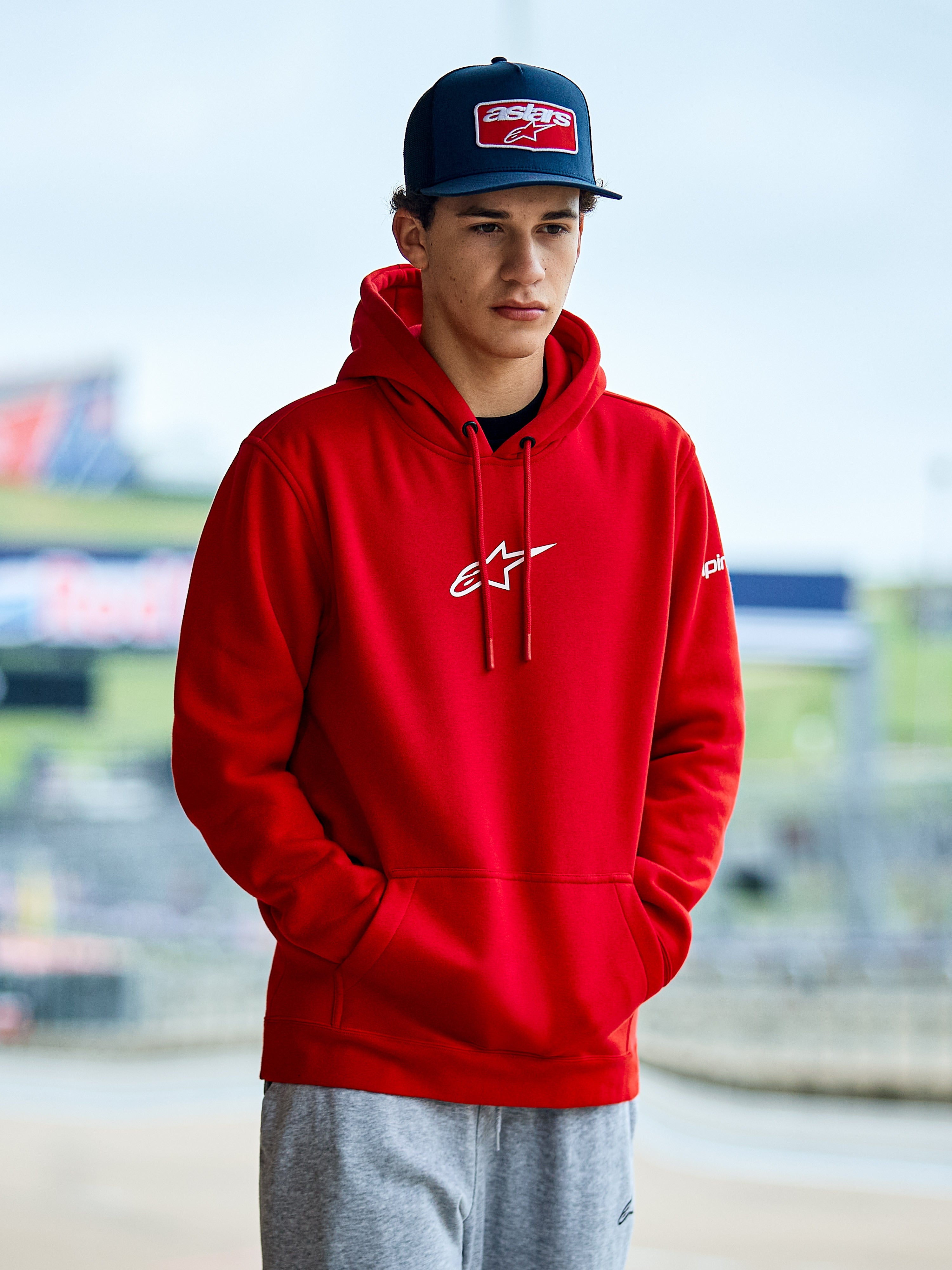 Alpinestars Frontal Hoodie, rode pullover fleece hoodie, voorzien van een wit Astars-logo op de borst, kangoeroezak en capuchon met trekkoord, gedragen door een man met een blauwe pet, casual motorsport streetwear