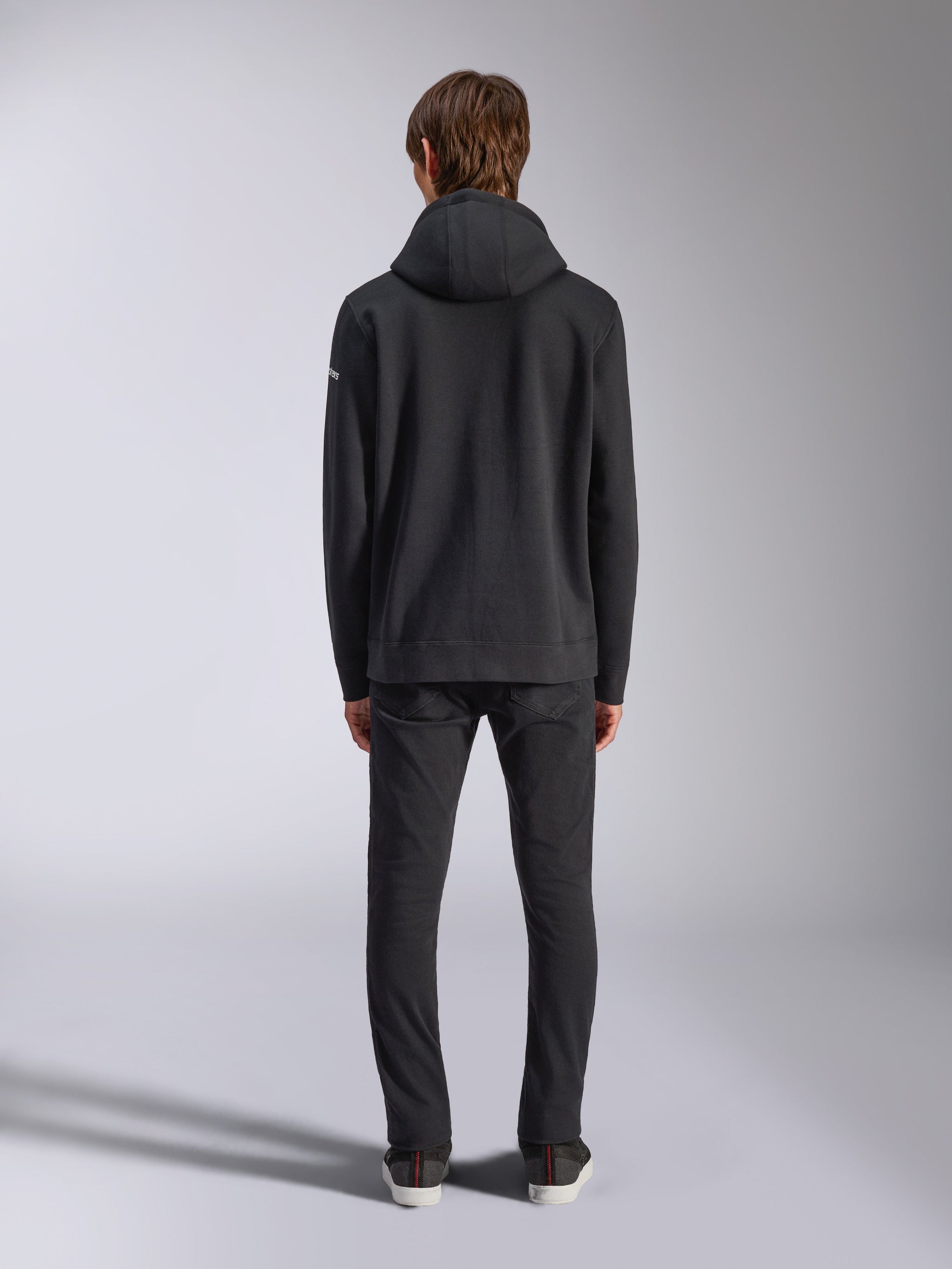 Alpinestars Frontal Hoodie, lässige Streetwear, Schwarz, Rückansicht eines Models im Fleece-Kapuzenpullover, mit Rippbündchen an Saum und Ärmeln, kleines weißes Wortmarken-Logo am Ärmel, kombiniert mit schwarzer Hose