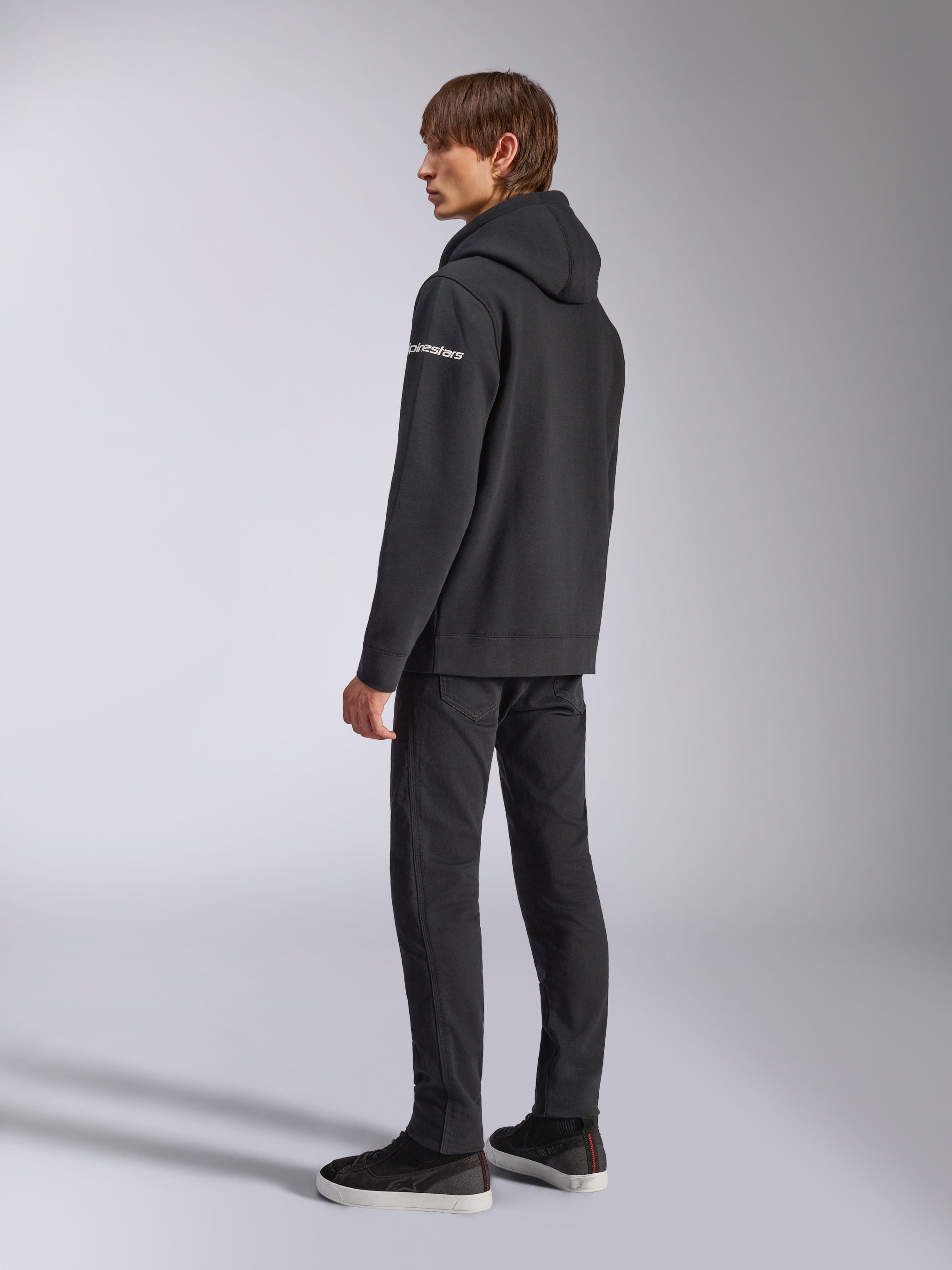 Alpinestars Frontal Hoodie, lässige Streetwear, Schwarz, Rückansicht von der Seite mit weißem Wortmarken-Logo am Ärmel, getragen mit schwarzer Hose und Sneakern, modernes motorsport-inspiriertes Design