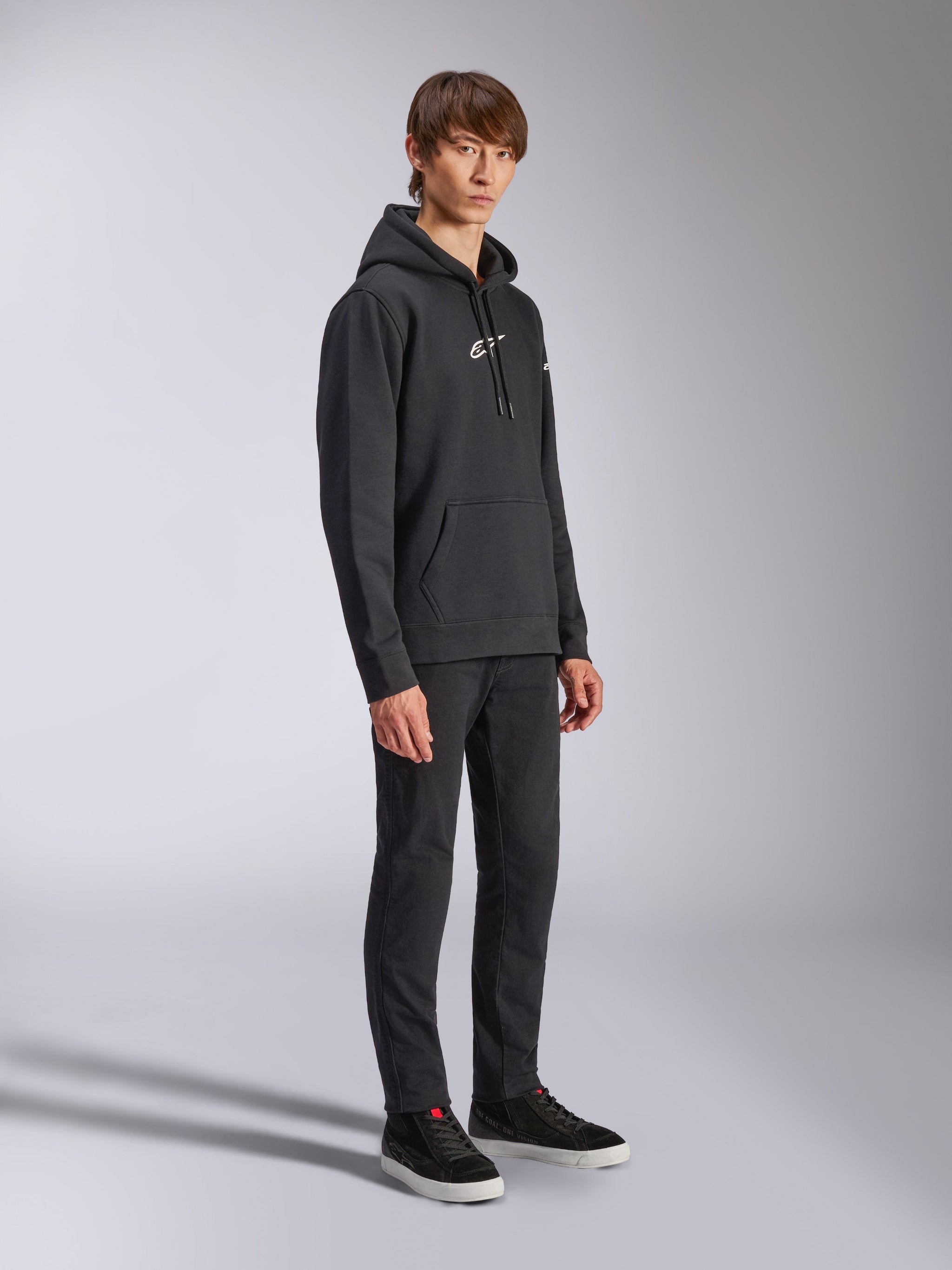 Alpinestars Frontal Hoodie, Schwarz, Fleece-Kapuzenpullover mit weißem HD-Logo auf der Brust und Wortmarken-Logos auf den Ärmeln, Kängurutasche, kombiniert mit schwarzer Hose und Sneakern, lässige vom Motorsport inspirierte Streetwear