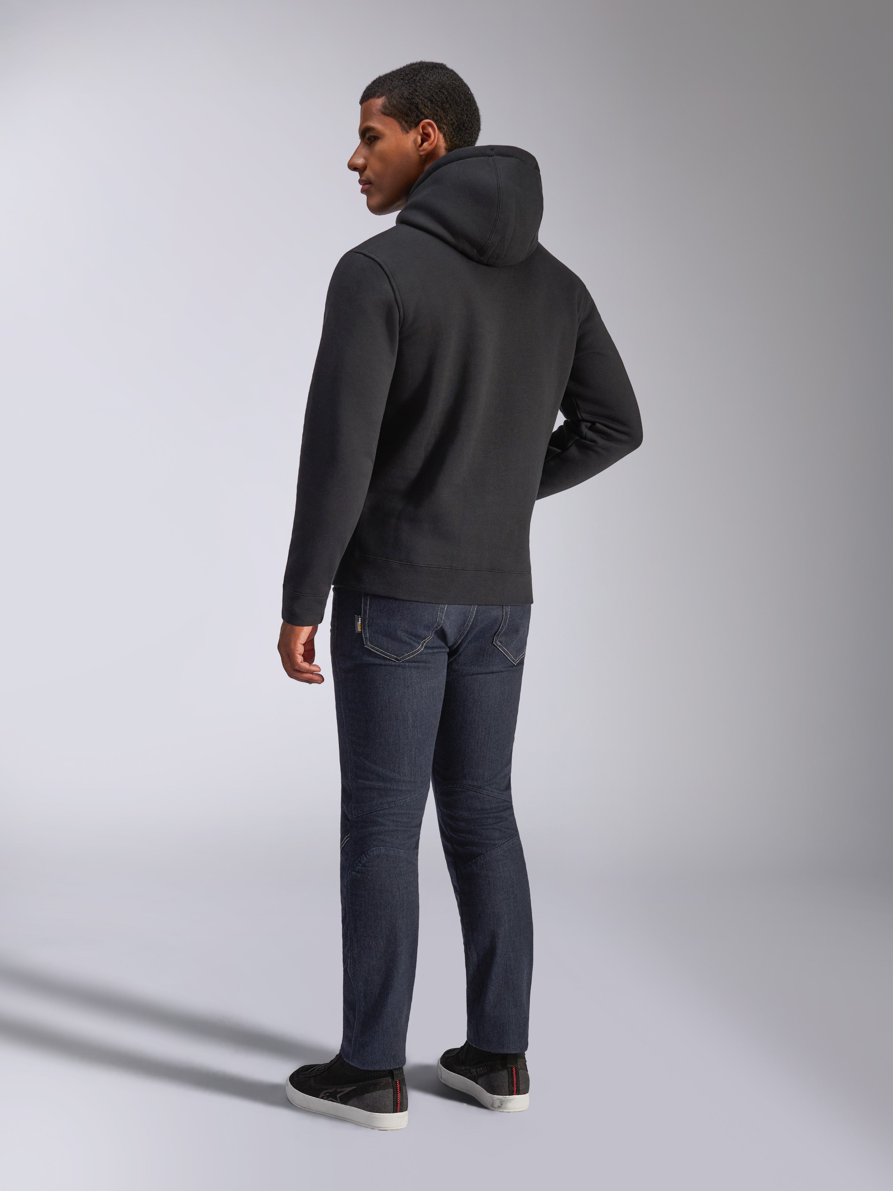 Alpinestars Blaze V3 Hoodie, back view of a man wearing the black pullover hoodie, paired with dark jeans and black sneakers, casual motorsport-inspired streetwear