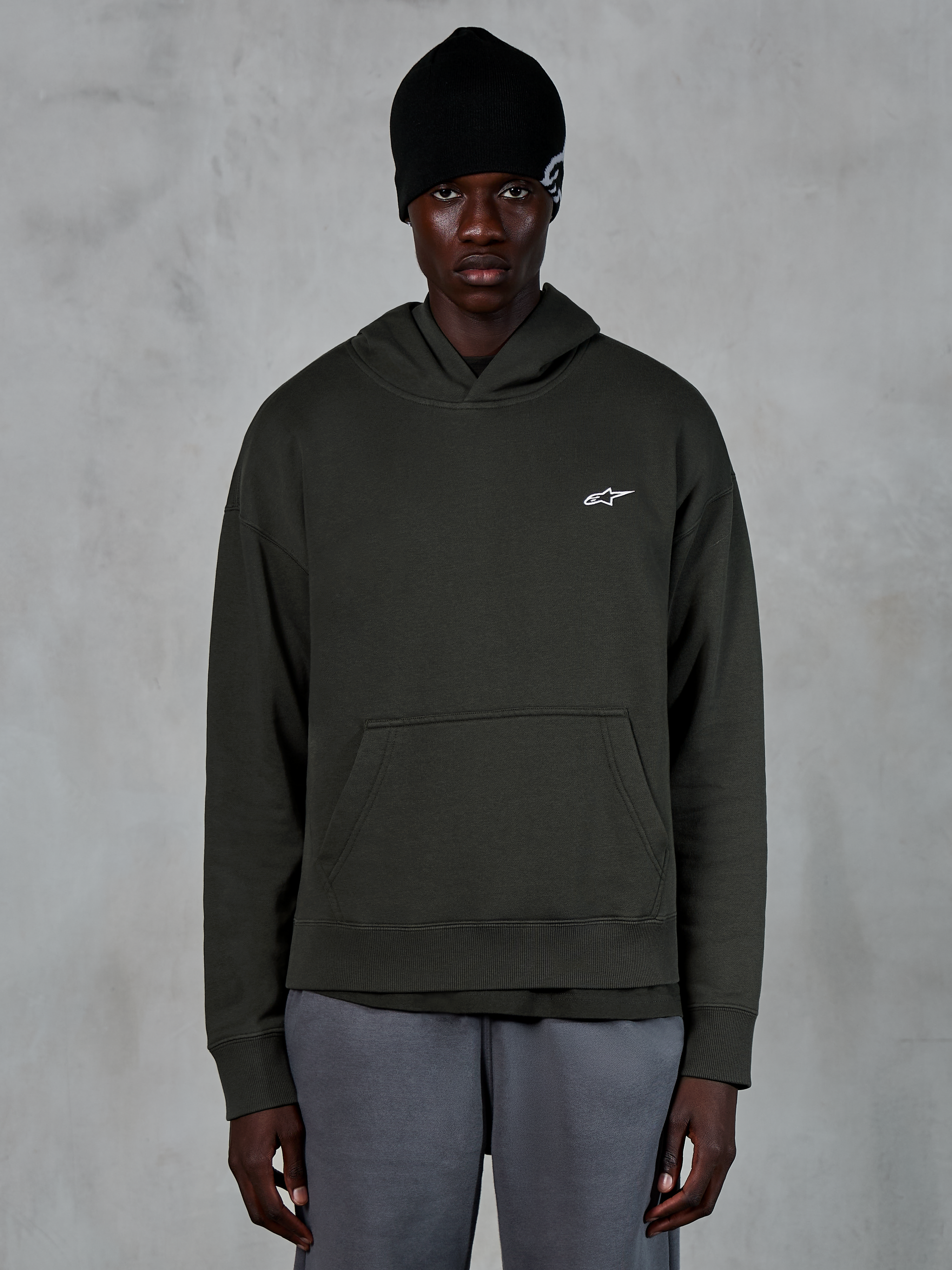 Alpinestars Essentials Ageless Hoodie, casual streetwear, Off Black pullover met een klein wit Alpinestars-logo op de borst, gedragen met een zwarte beanie, minimalistisch ontwerp met een kangoeroezak aan de voorzijde en stone-wash afwerking