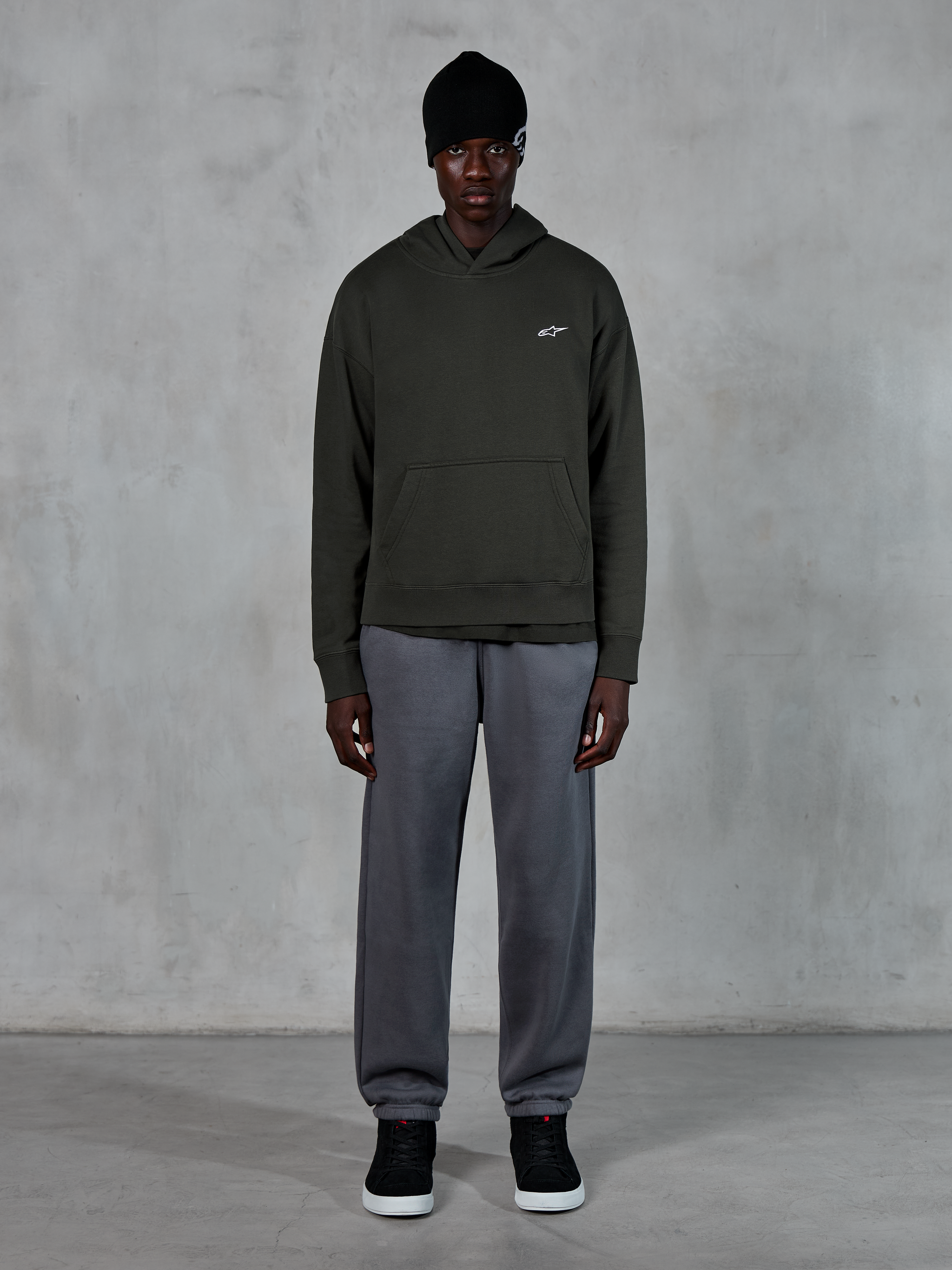 Model draagt Alpinestars Essentials Ageless Hoodie, casual streetwear, Off Black met klein wit logo op de borst, gecombineerd met grijze joggingbroek, zwarte beanie en zwarte sneakers, minimalistisch op motorsport geïnspireerd design