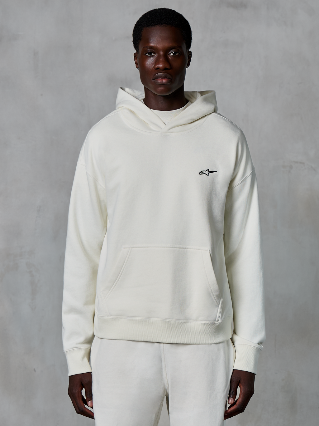 Alpinestars Essentials Ageless Hoodie, off-white, worn by male model, small black logo on chest, front pouch pocket, long sleeves, casual motorsport-inspired streetwear
