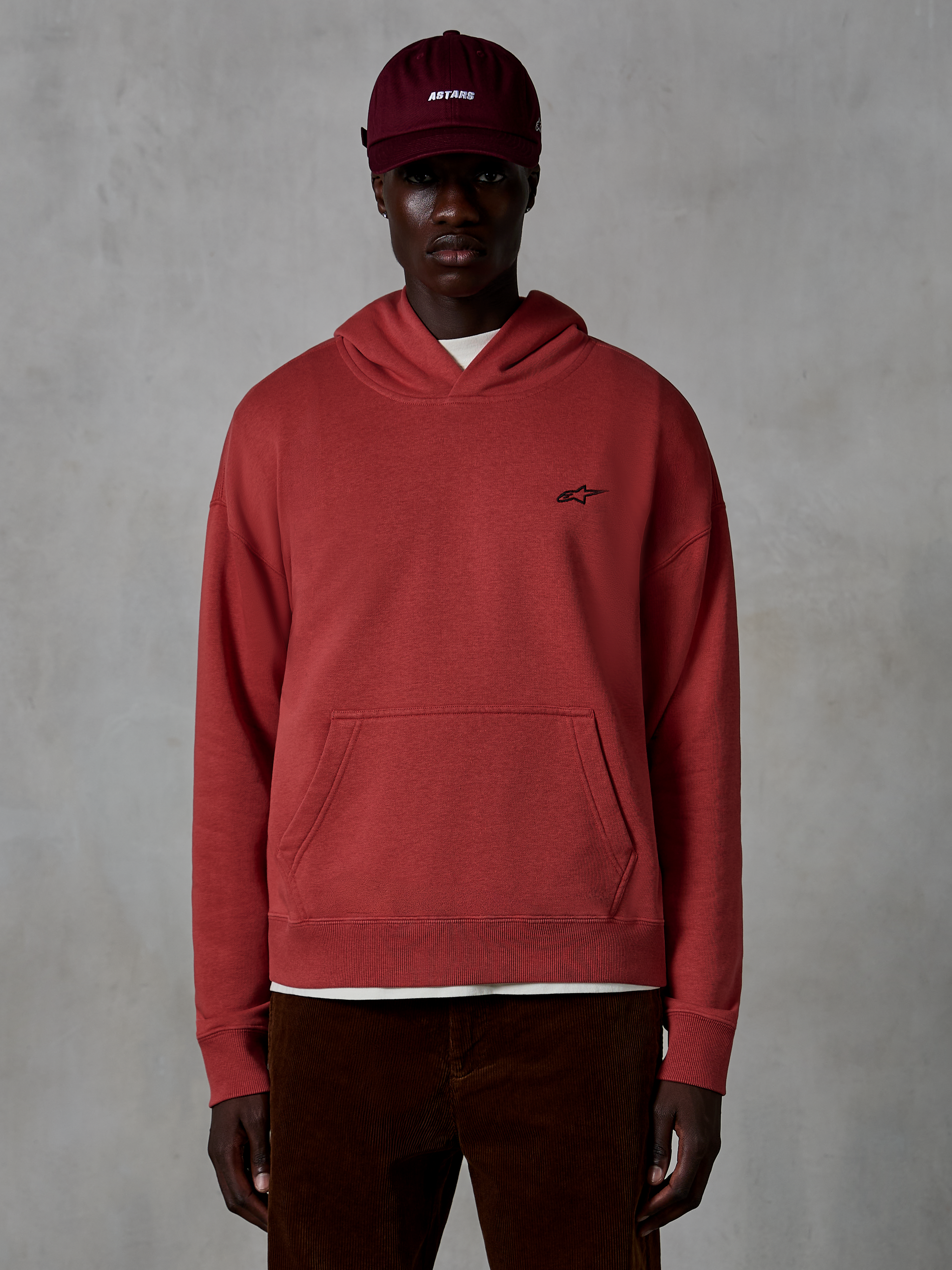 Alpinestars Essentials Ageless Hoodie, casual streetwear, steenrode kleur, voorzien van een klein zwart sterlogo op de borst, kangoeroezak, stonewash afwerking, gedragen door een model met een bordeauxrode pet en bruine corduroy broek.