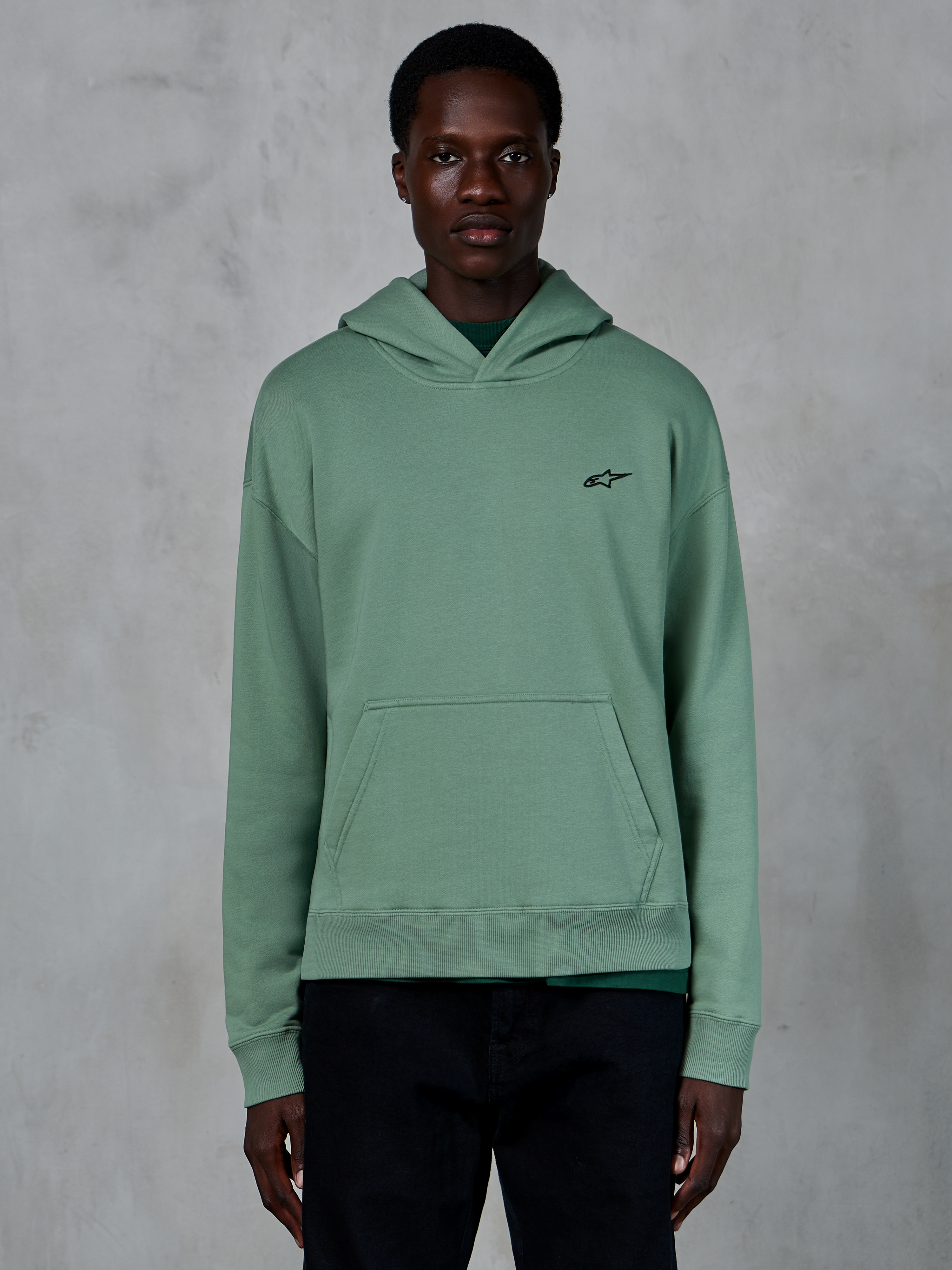 Alpinestars Essentials Ageless Hoodie, Kaktusgrün, lässiger Fleece-Hoodie mit Stone-Wash-Finish, kleines schwarzes Stern-Logo auf der Brust, Kängurutasche vorne, getragen von einem Model in einem schlichten Lifestyle-Umfeld