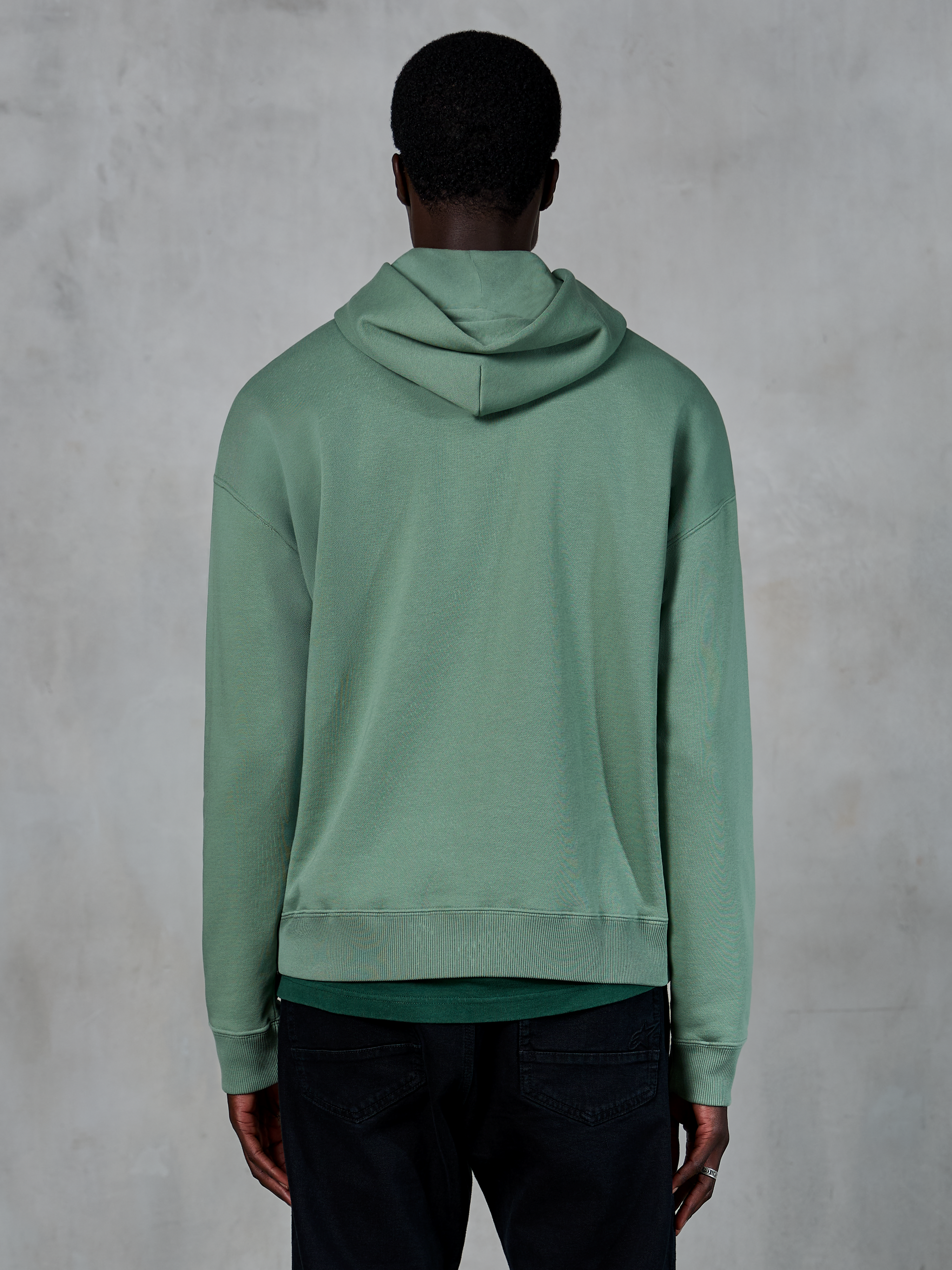 Alpinestars Essentials Ageless Hoodie, lässige Sportbekleidung, Kaktusgrün, Rückansicht am Modell, Garment-Stone-Wash-Finish, kombiniert mit schwarzer Hose, minimalistisches Lifestyle-Design