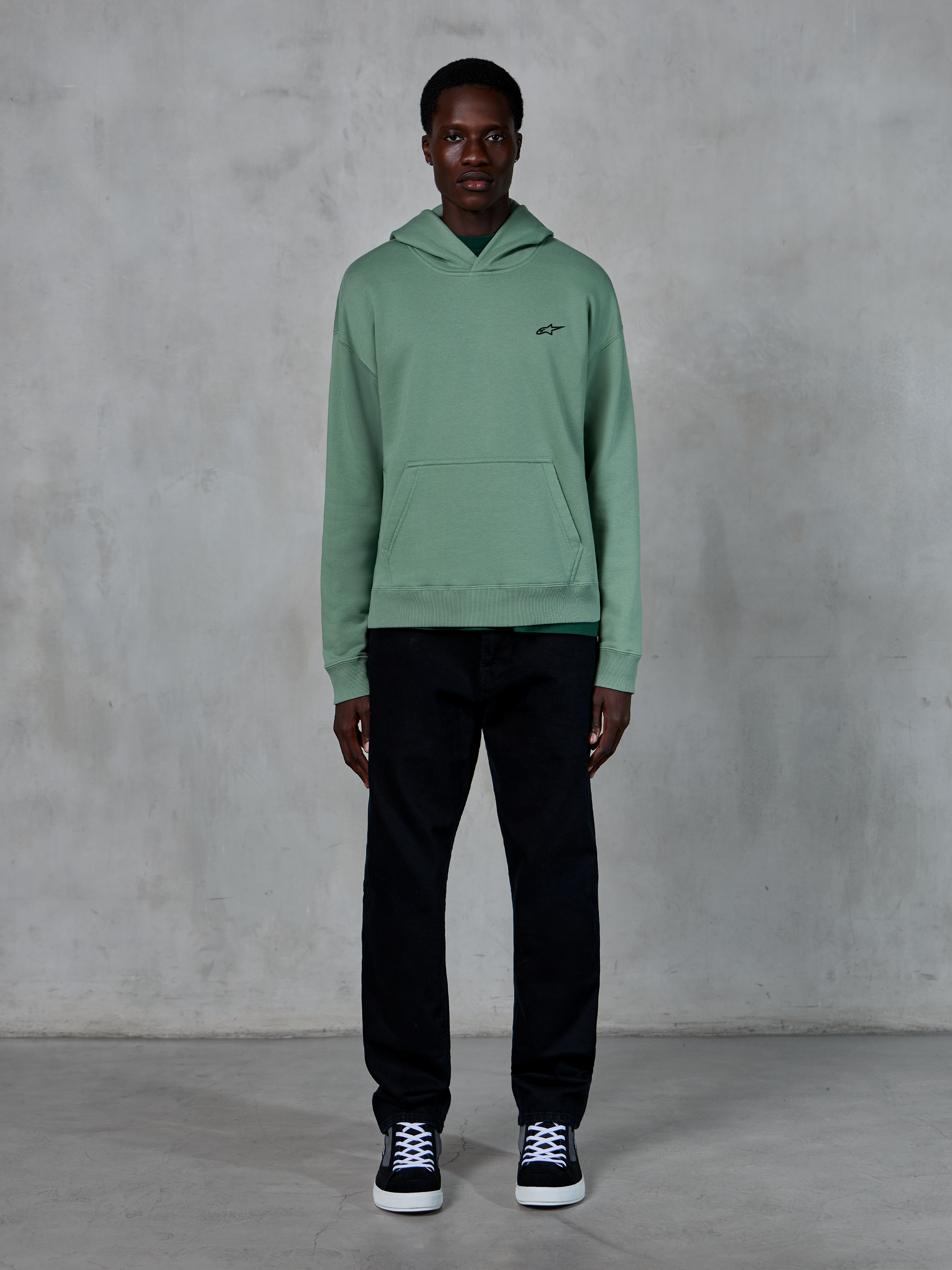 Alpinestars Essentials Ageless Hoodie, Kaktusgrün, kleines schwarzes Brustlogo, Kängurutasche, lässige Lifestyle-Streetwear getragen mit schwarzer Hose und schwarz-weißen Sneakern vor einer strukturierten Wand