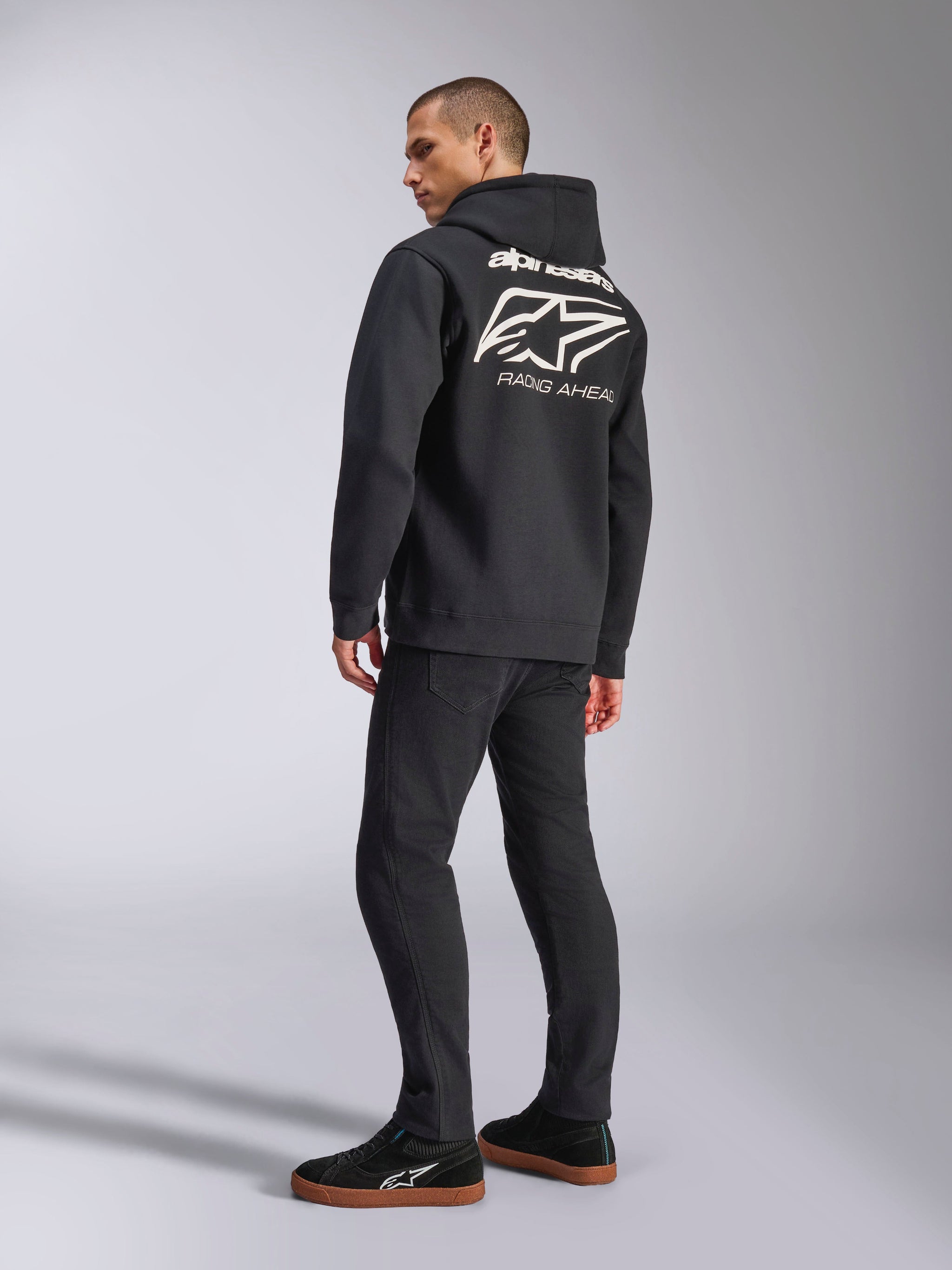 Model im Alpinestars Formulation Hoodie, Schwarz, Rückansicht mit großem weißem Alpinestars-Logo und „RACING AHEAD“-Schriftzug, lässige motorsport-inspirierte Streetwear, kombiniert mit schwarzer Hose und schwarzen Sneakern