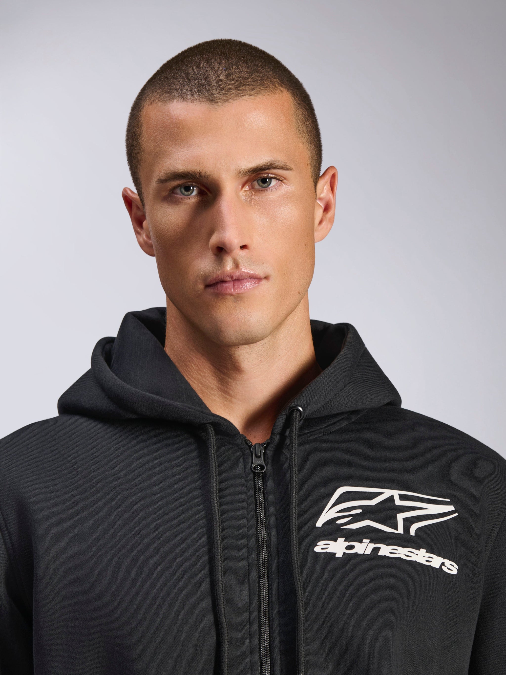 Alpinestars Formulation Hoodie, Schwarz, männliches Model trägt Poly-Fleecejacke mit Reißverschluss, weißes Alpinestars-Logo auf der Brust, Kapuze mit Kordelzug, lässige, vom Motorsport inspirierte Streetwear