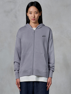 Alpinestars Essentials Ageless Zip Hoodie, Silver, casual streetwear, fleece sweatshirt met volledige ritssluiting en een klein zwart Alpinestars-logo op de borst, gedeelde kangoeroezak, gedragen door model over een wit t-shirt en donkere jeans