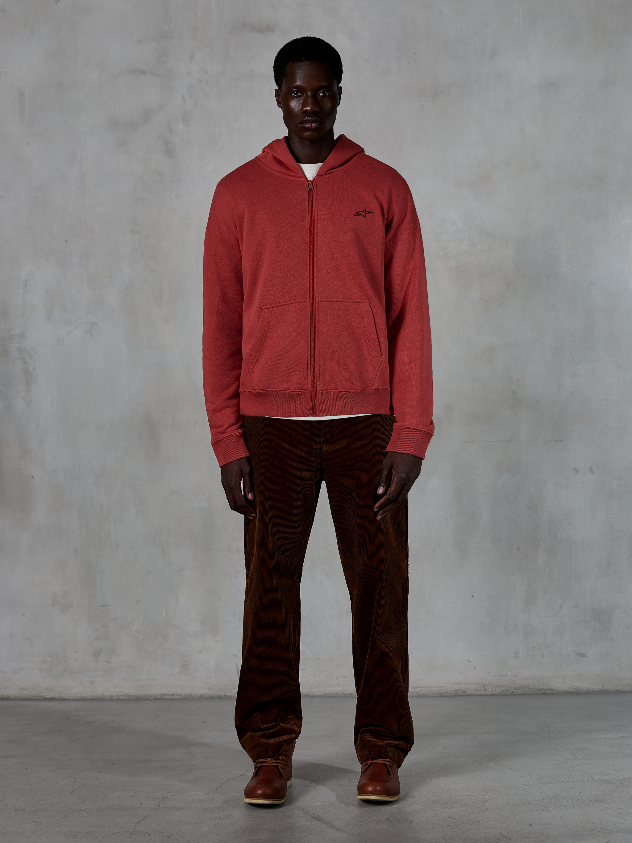 Alpinestars Essentials Ageless Zip Hoodie, lässige Streetwear, ziegelrot mit kleinem schwarzem Sternenlogo, durchgehender Reißverschluss vorne, getragen von einem Model mit brauner Cordhose und Stiefeln, hochwertiges, vom Motorsport inspiriertes Design