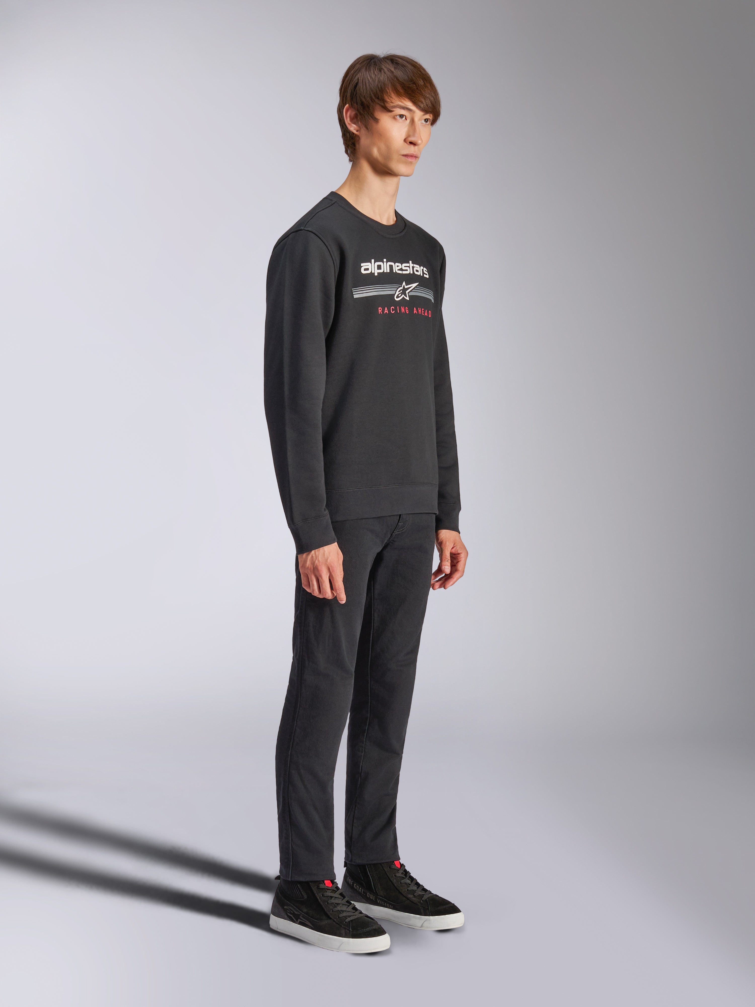 Sweatshirt Alpinestars Bettern Crew, pull à col rond noir, logo Alpinestars blanc avec l'inscription 'RACING AHEAD' en rouge sur la poitrine, associé à un pantalon noir et des baskets, style streetwear décontracté inspiré du sport automobile