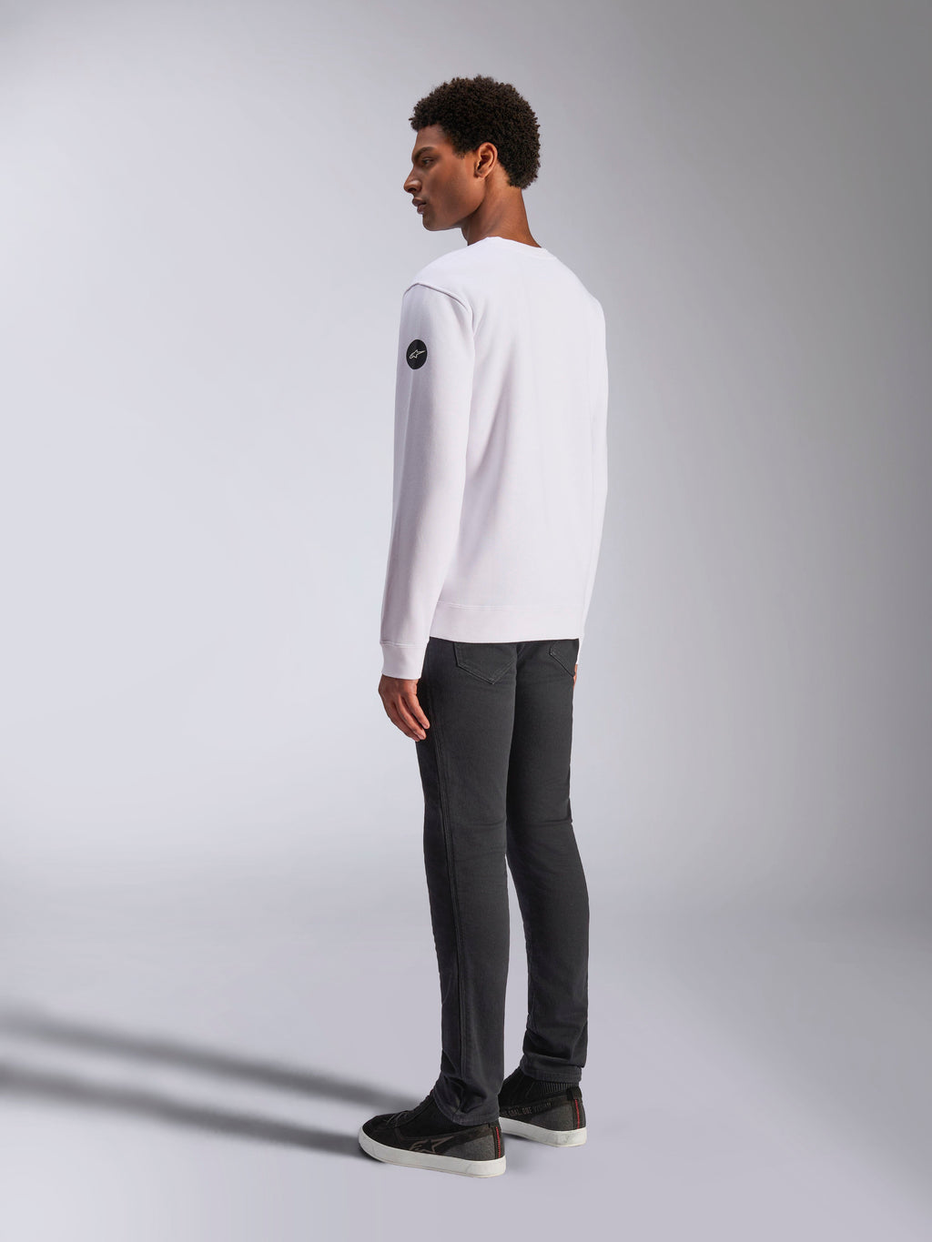 Alpinestars Bettern Crew, sweat-shirt à col rond blanc, vue de dos et de côté, logo circulaire noir sur la manche, porté avec un pantalon gris foncé et des baskets noires, style streetwear motorsport décontracté