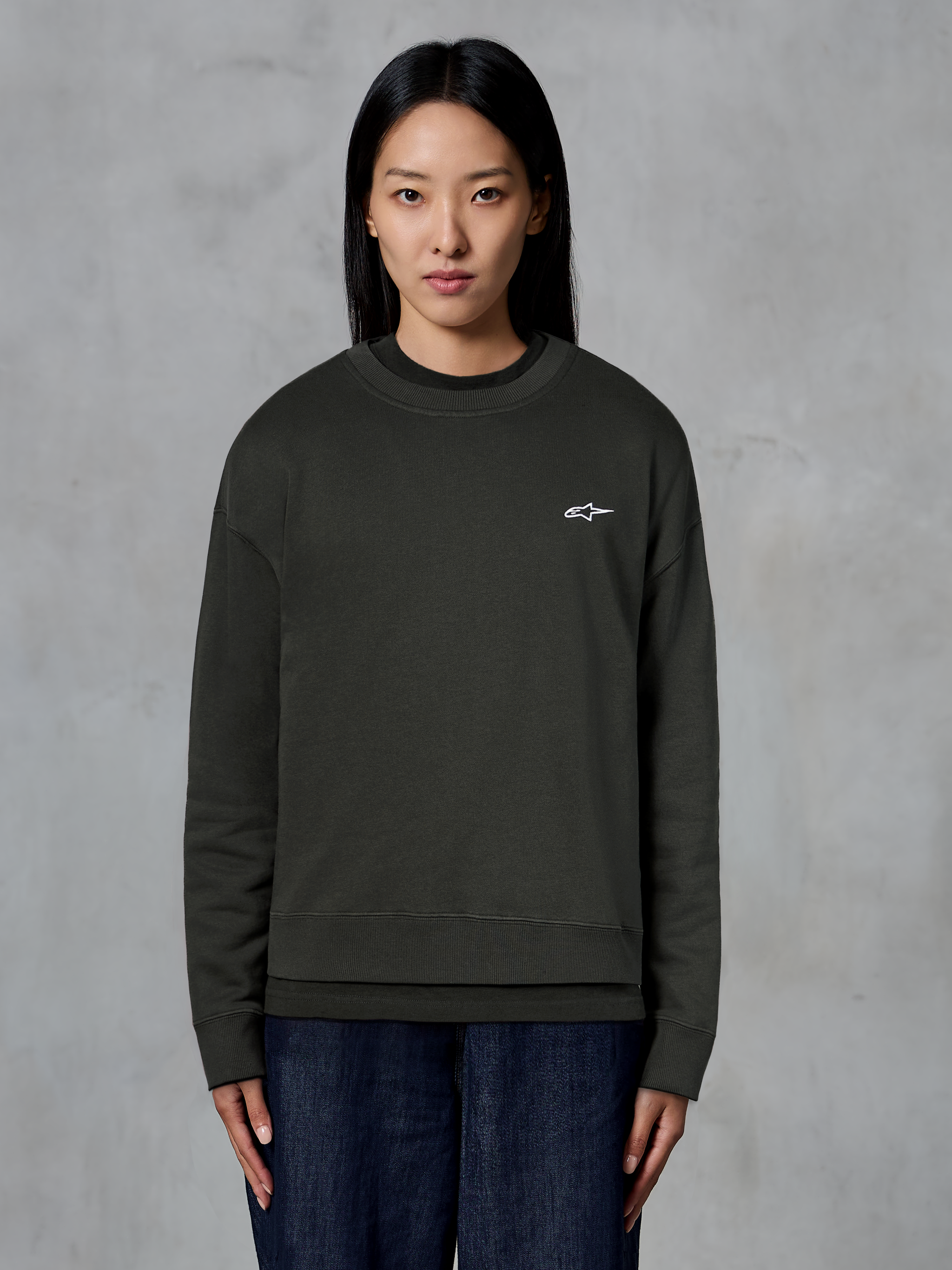 Alpinestars Essentials Ageless Crew, hochwertiges Rundhals-Sweatshirt, Off Black, mit einem kleinen weißen Alpinestars-Logo auf der Brust, lässiger Streetwear-Stil, getragen von einem Model mit dunkler Jeans