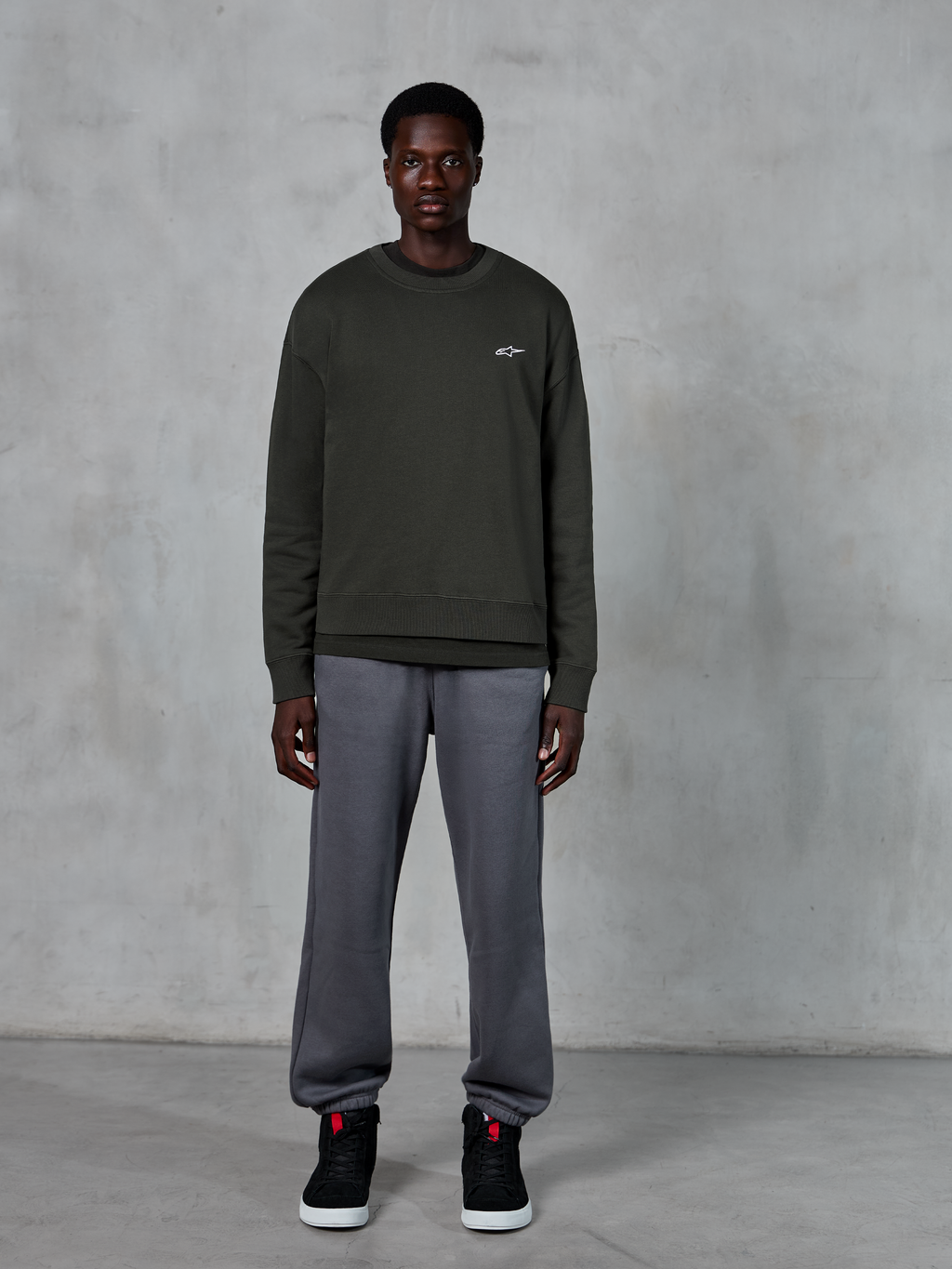 Alpinestars Essentials Ageless Crew, dunkelgrünes Rundhals-Sweatshirt mit kleinem weißem Logo auf der Brust, getragen von einem Model mit grauer Jogginghose und schwarzen High-Top-Sneakern, lässige Lifestyle-Bekleidung